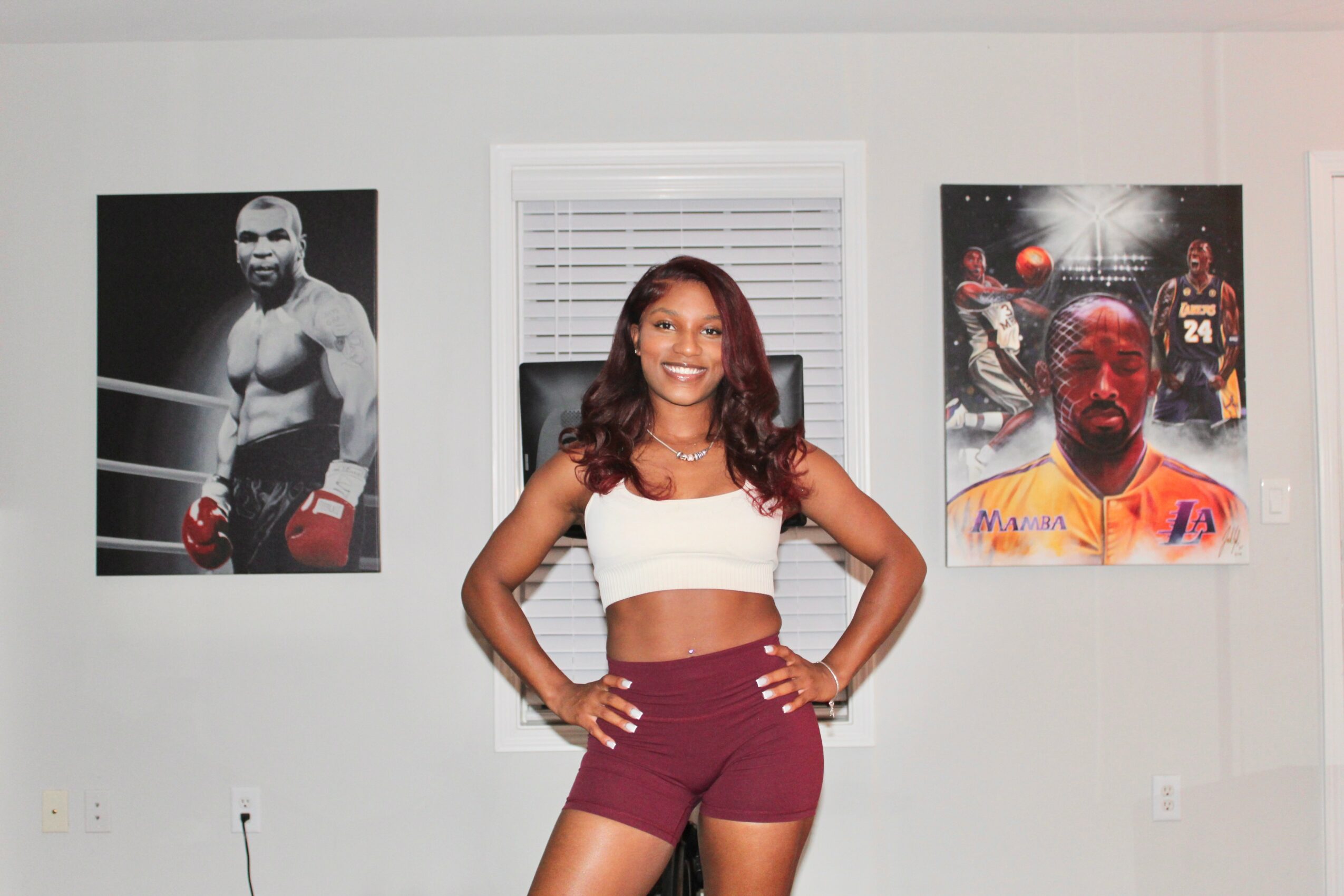 Woman with long hair in a white crop top and maroon shorts smiling with hands on hips, standing in front of a window.