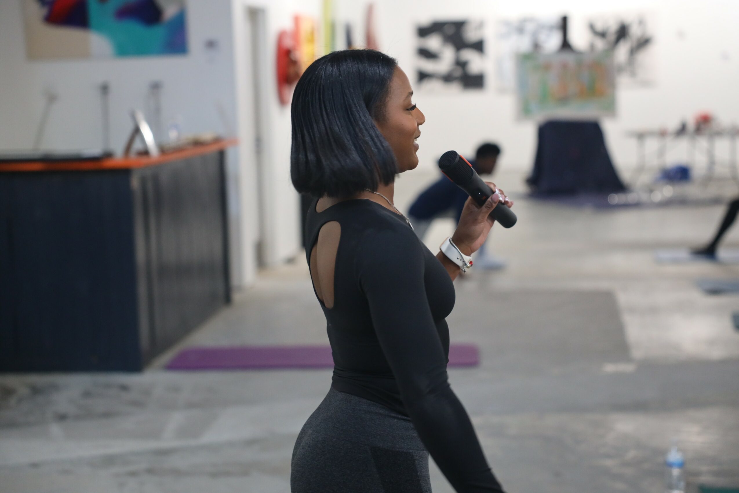 Woman with black hair holding a microphone, standing indoors with blurred background, facing right.