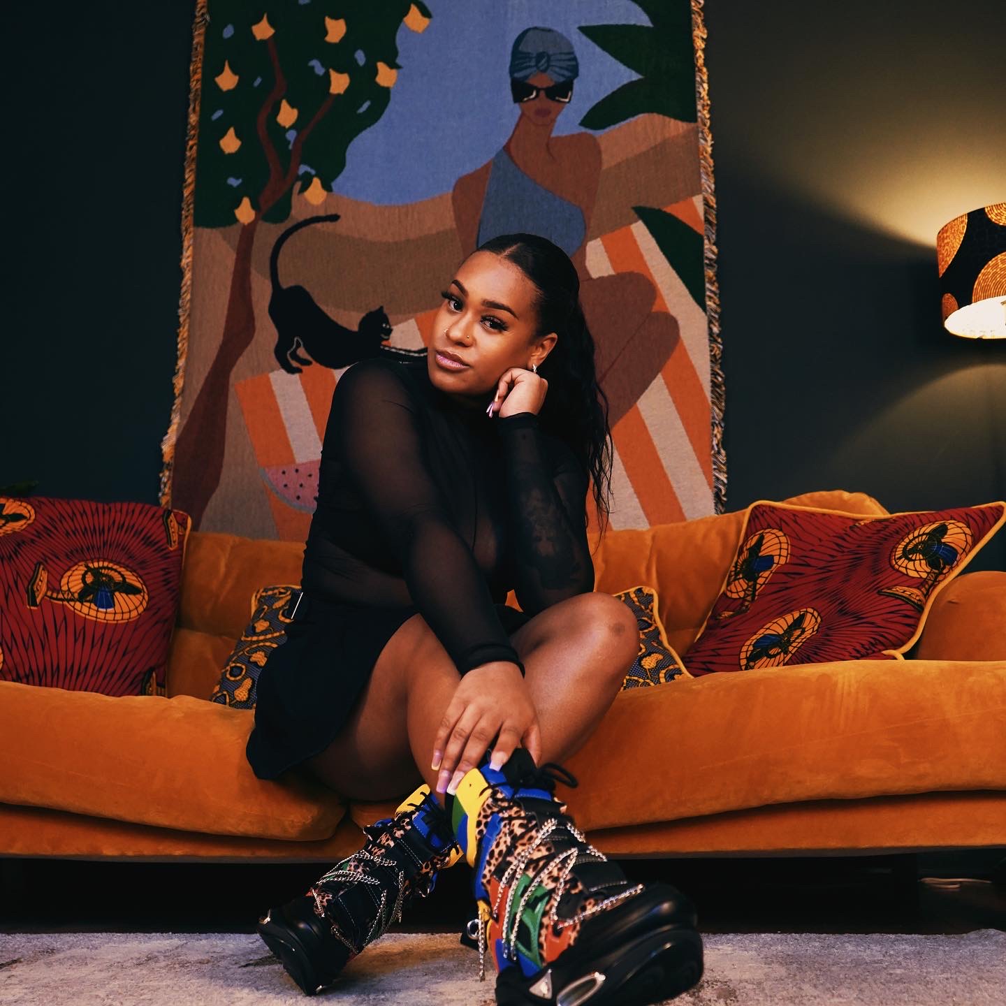 Young woman sitting on a mustard-colored sofa with colorful pillows, in a room with a vibrant wall hanging and dim lighting.
