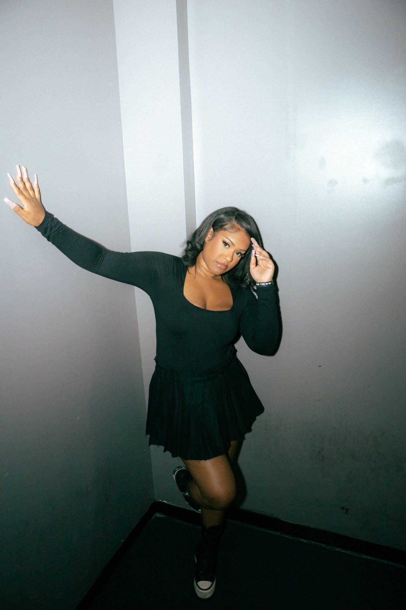 Woman with long dark hair in black dress and stockings, posing with one arm raised, in a corner with gray walls.