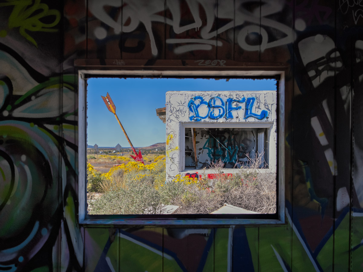 A window with graffiti on a wall, overlooking a landscape with a large arrow sculpture and yellow flowers.