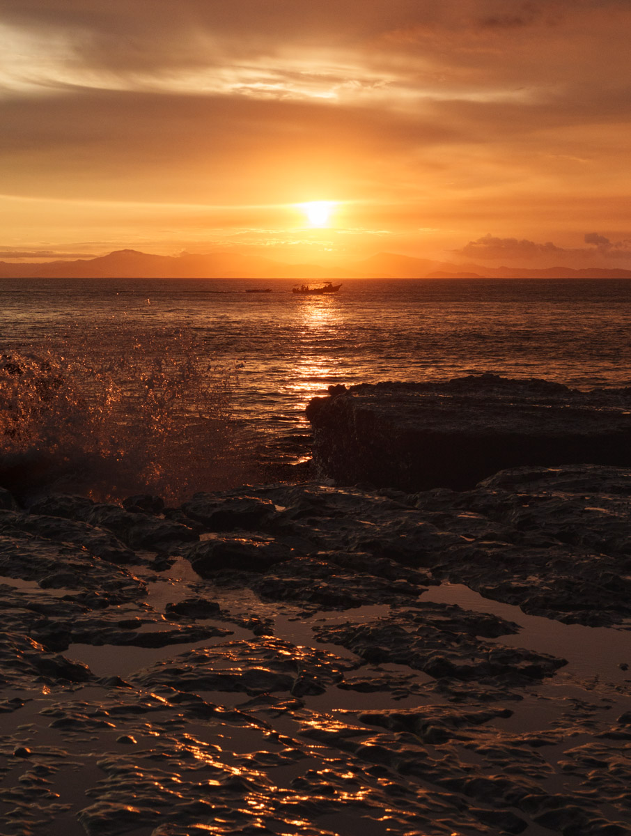 Sunset over ocean with rocky shoreline, warm colors in sky, reflection on water, and distant land silhouette.