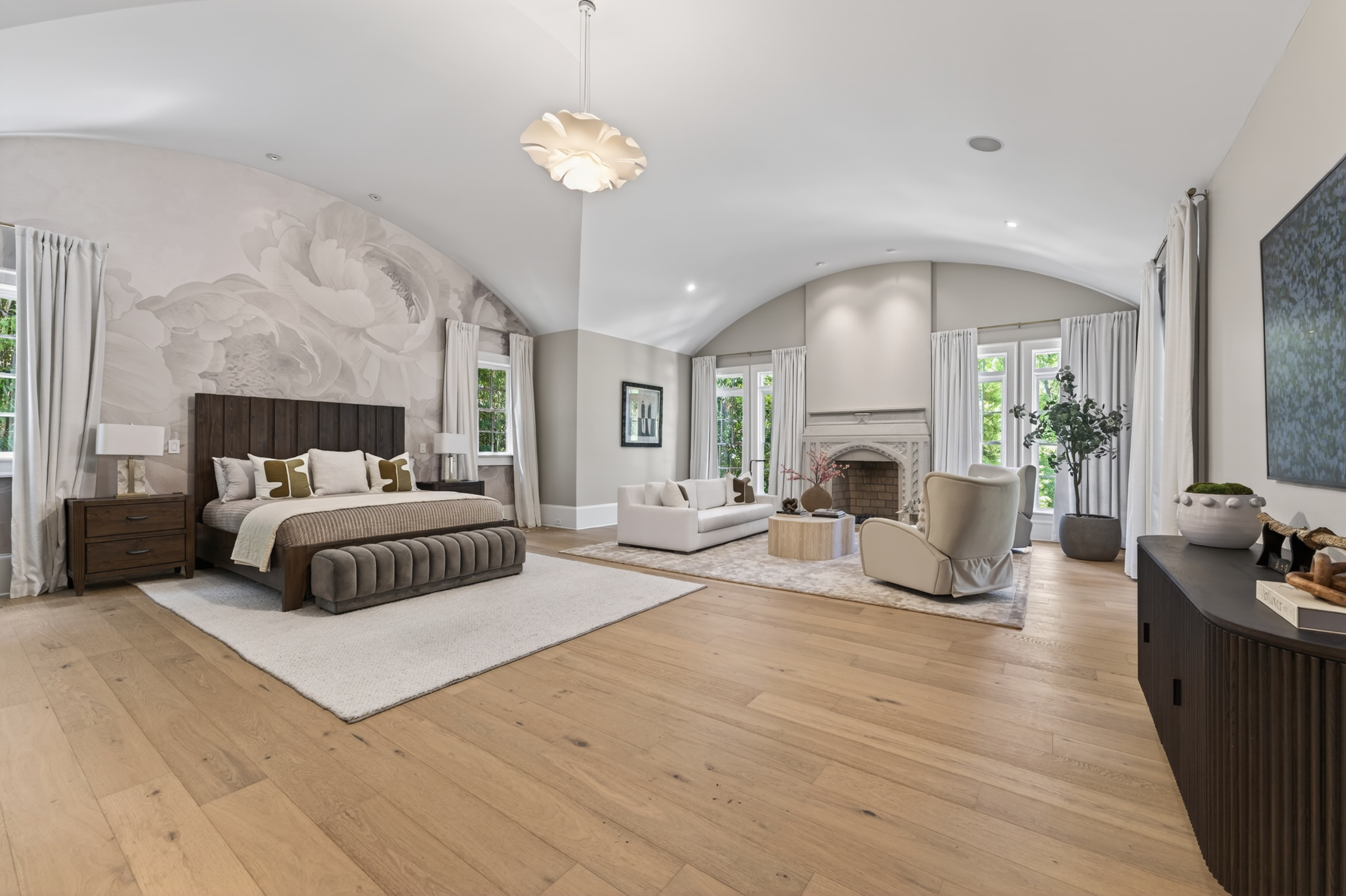 Spacious bedroom with a large bed, sitting area, fireplace, and large windows, bright and airy with wooden flooring.