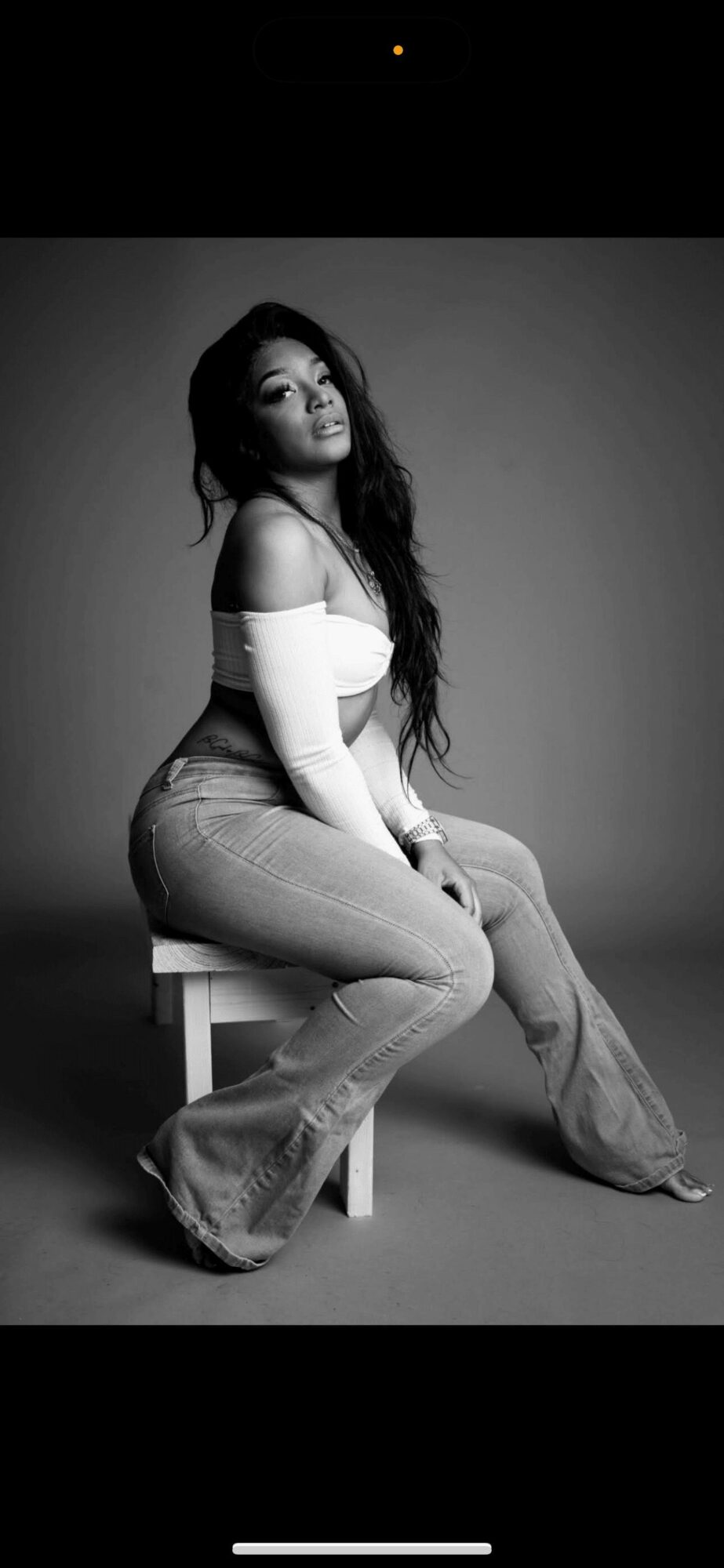 Woman with long hair sitting on a stool, wearing an off-shoulder top and jeans, looking at the camera.