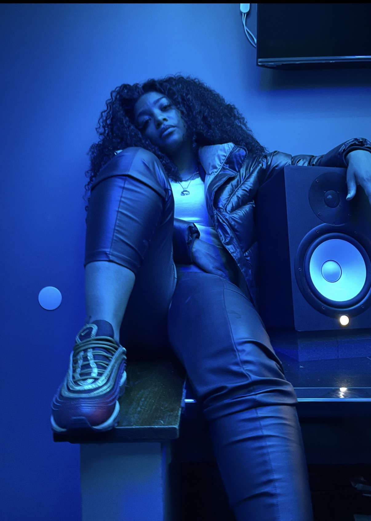 Person with curly hair sitting on a table in a dimly lit room with blue lighting, next to a speaker.