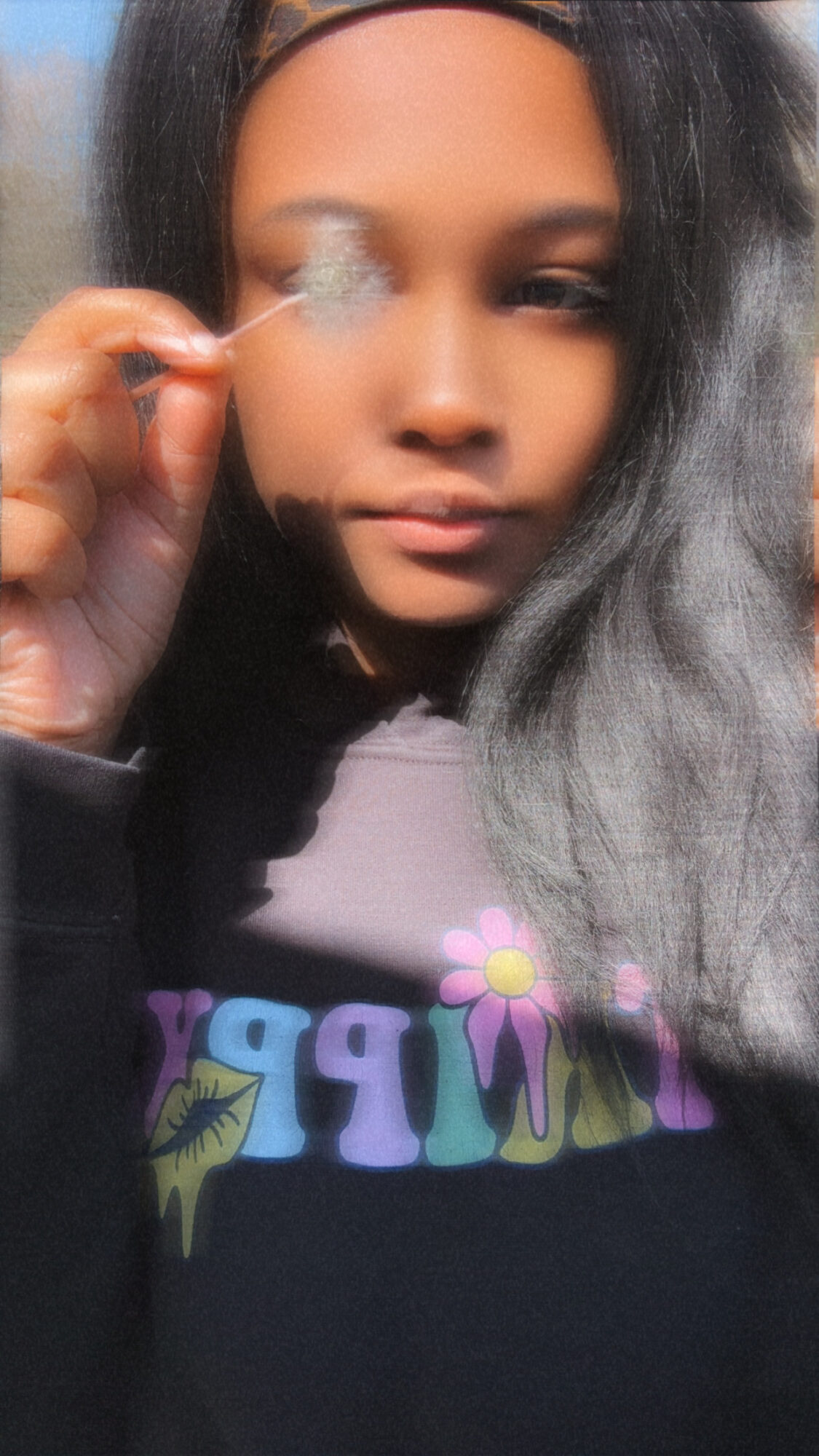 Young woman with long dark hair holding a small object near her face, wearing a black shirt with colorful text and a flower, neutral expression.