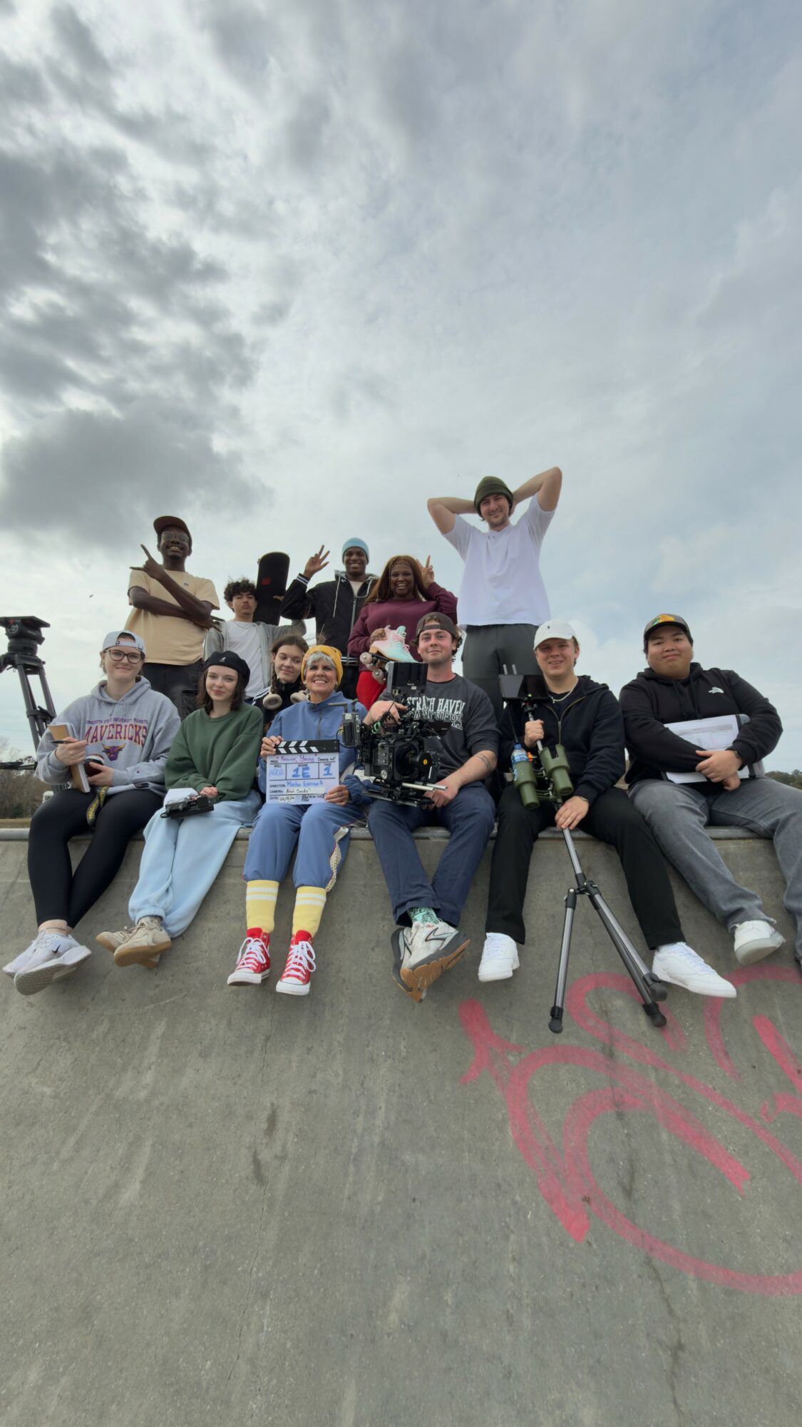 Group of ten people sitting and standing outdoors on a concrete surface with cloudy sky background.