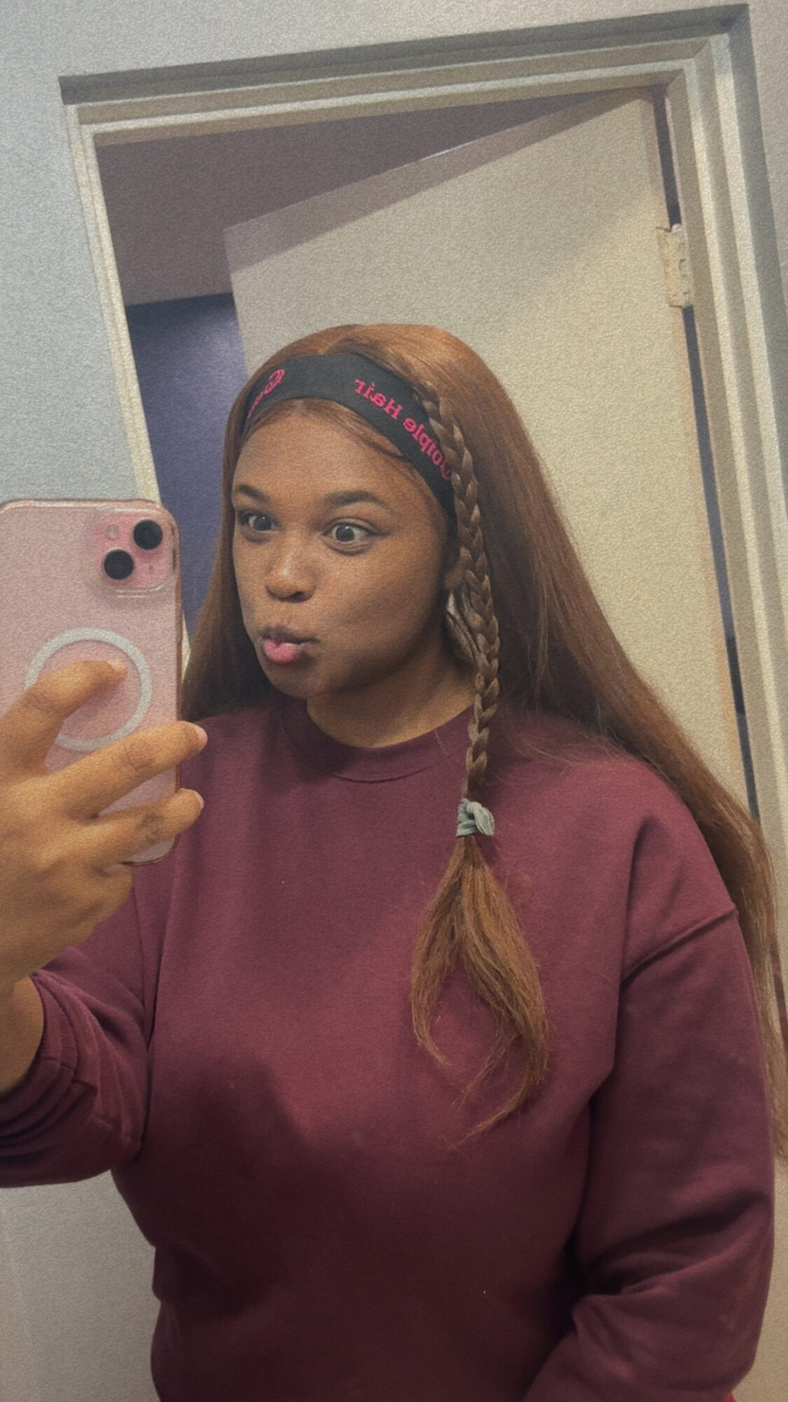 Young woman taking a mirror selfie with a pink phone, wearing a maroon sweatshirt and a headband, making a pout face.