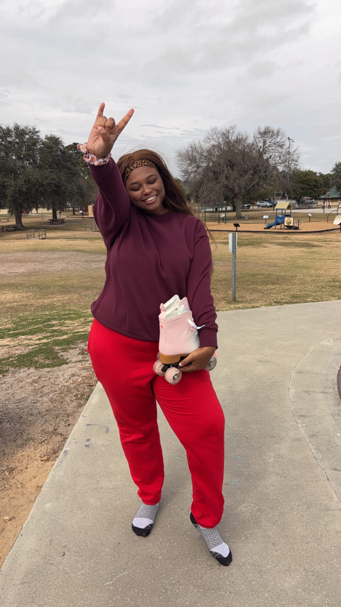 Person standing outdoors on a sidewalk, smiling, wearing a purple sweater and red pants, holding a small bag, making a hand gesture.