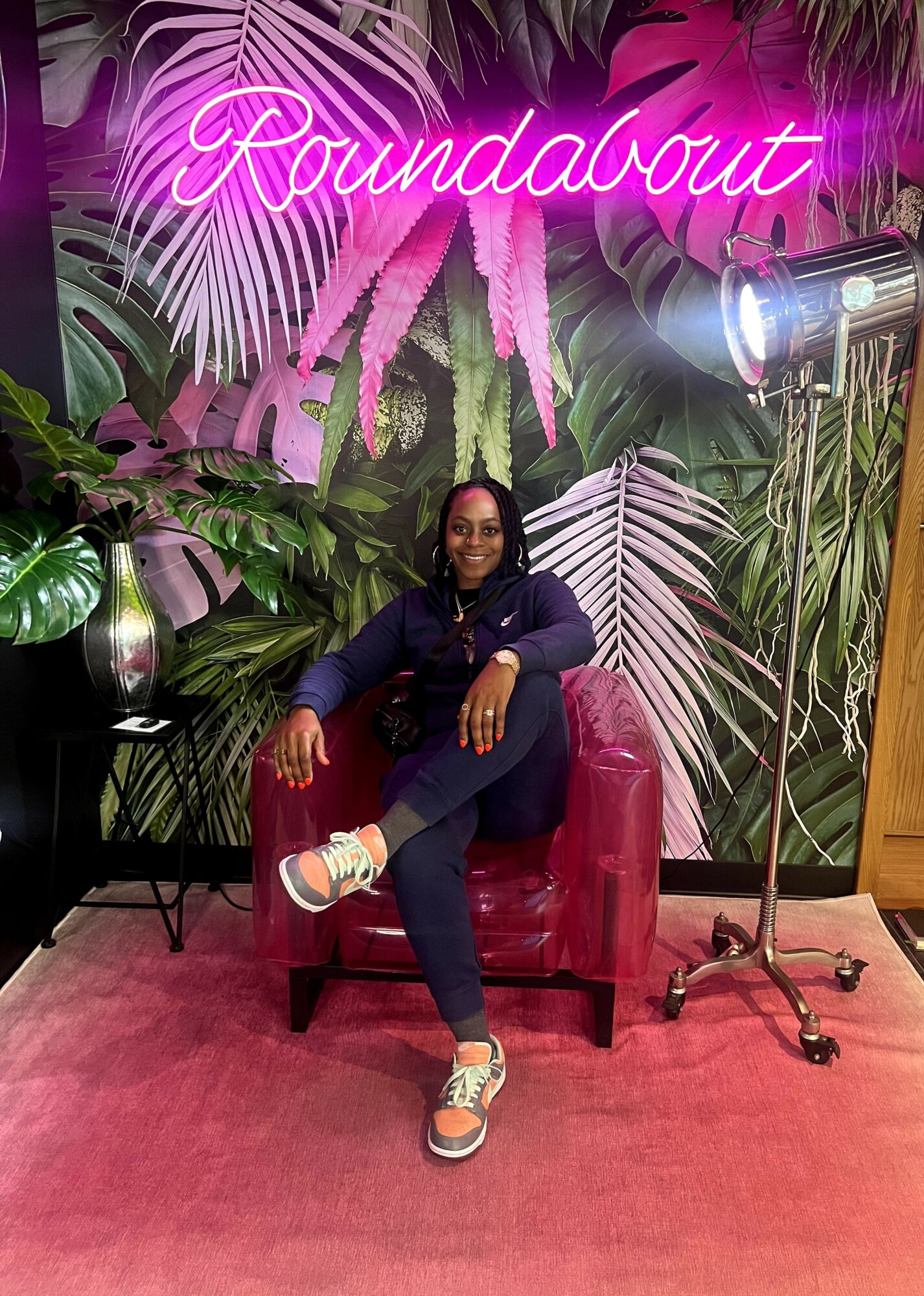 Woman sitting on a pink chair in front of a tropical plant backdrop with a neon sign reading 'Roundabout'.