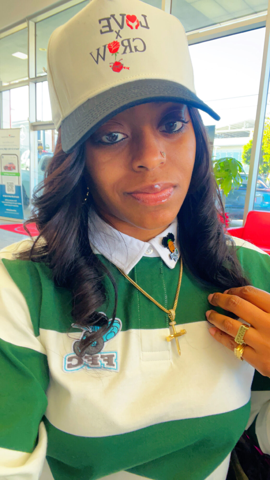 Young woman wearing a white cap with red and black text, green and white striped shirt, and gold jewelry, indoors.