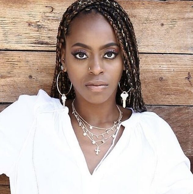 Woman with braided hair, wearing jewelry, against wooden background, looking directly at camera.