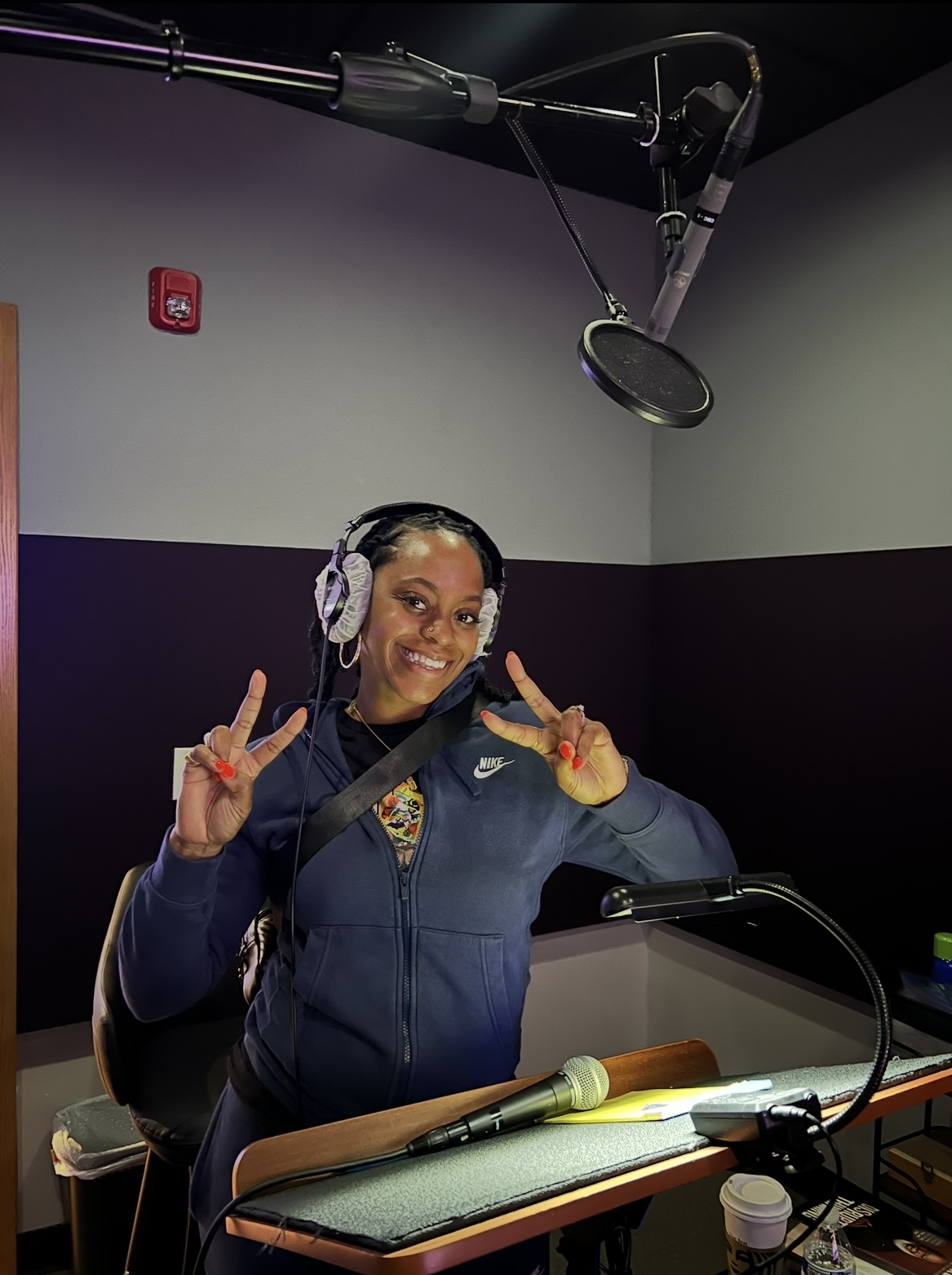 Woman in a recording studio wearing headphones, smiling, making peace signs, with microphone and recording equipment.