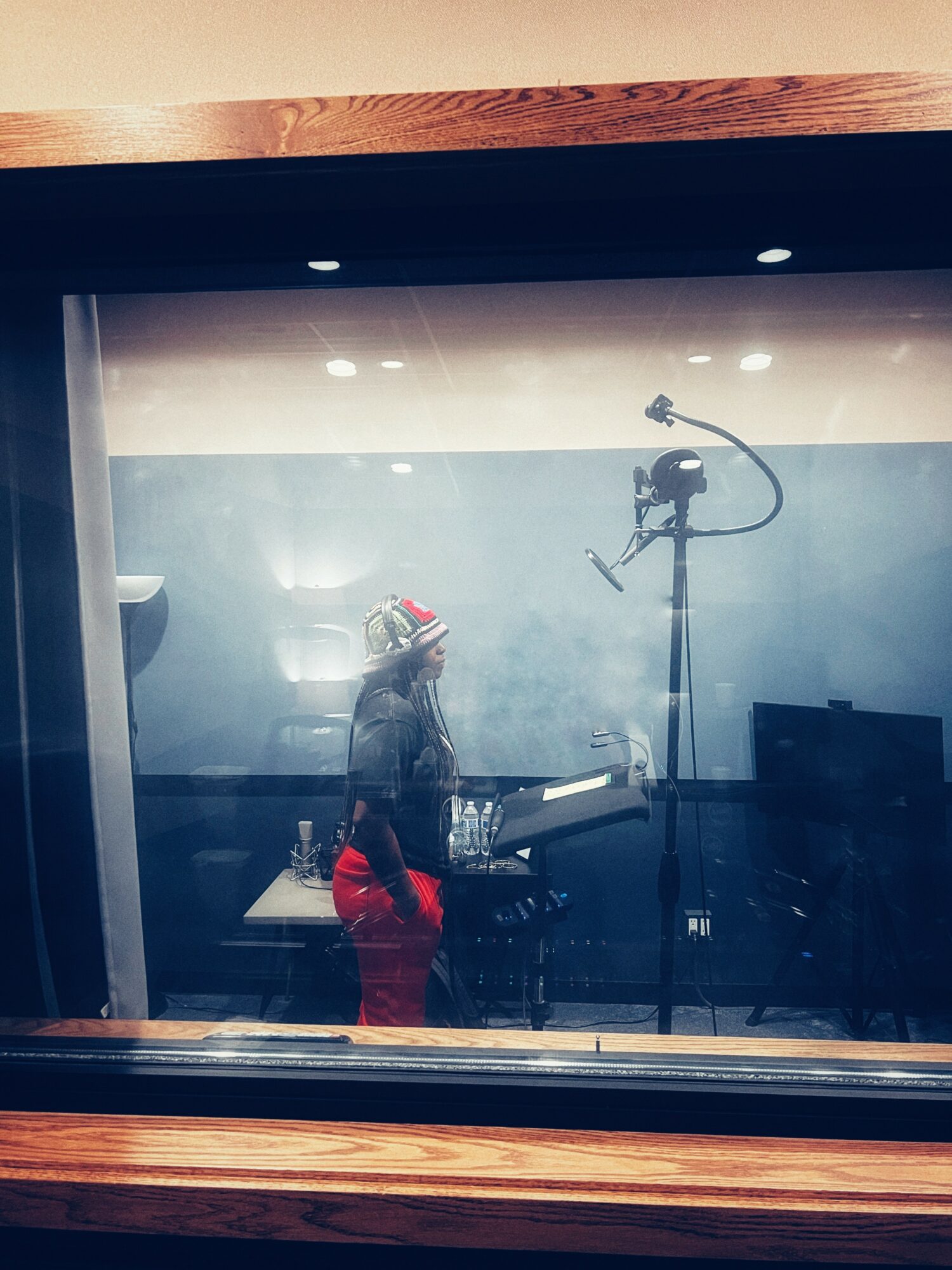 Person wearing a hat and red pants stands in a recording studio with equipment, seen through a glass window.
