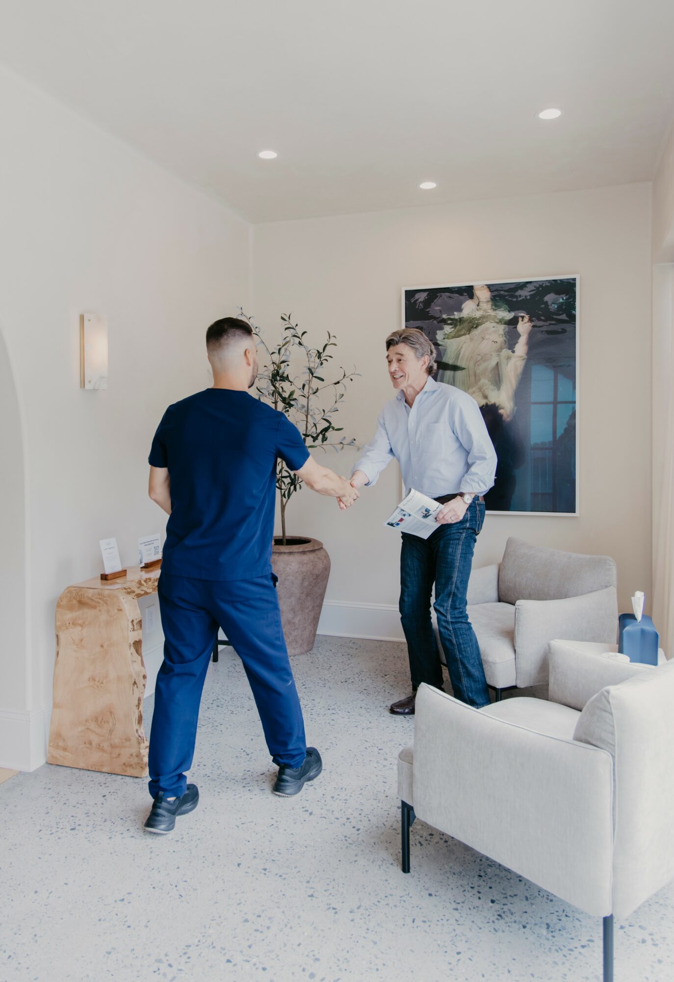 Two men shake hands in a modern, well-lit room with a large painting, a potted plant, and seating area.