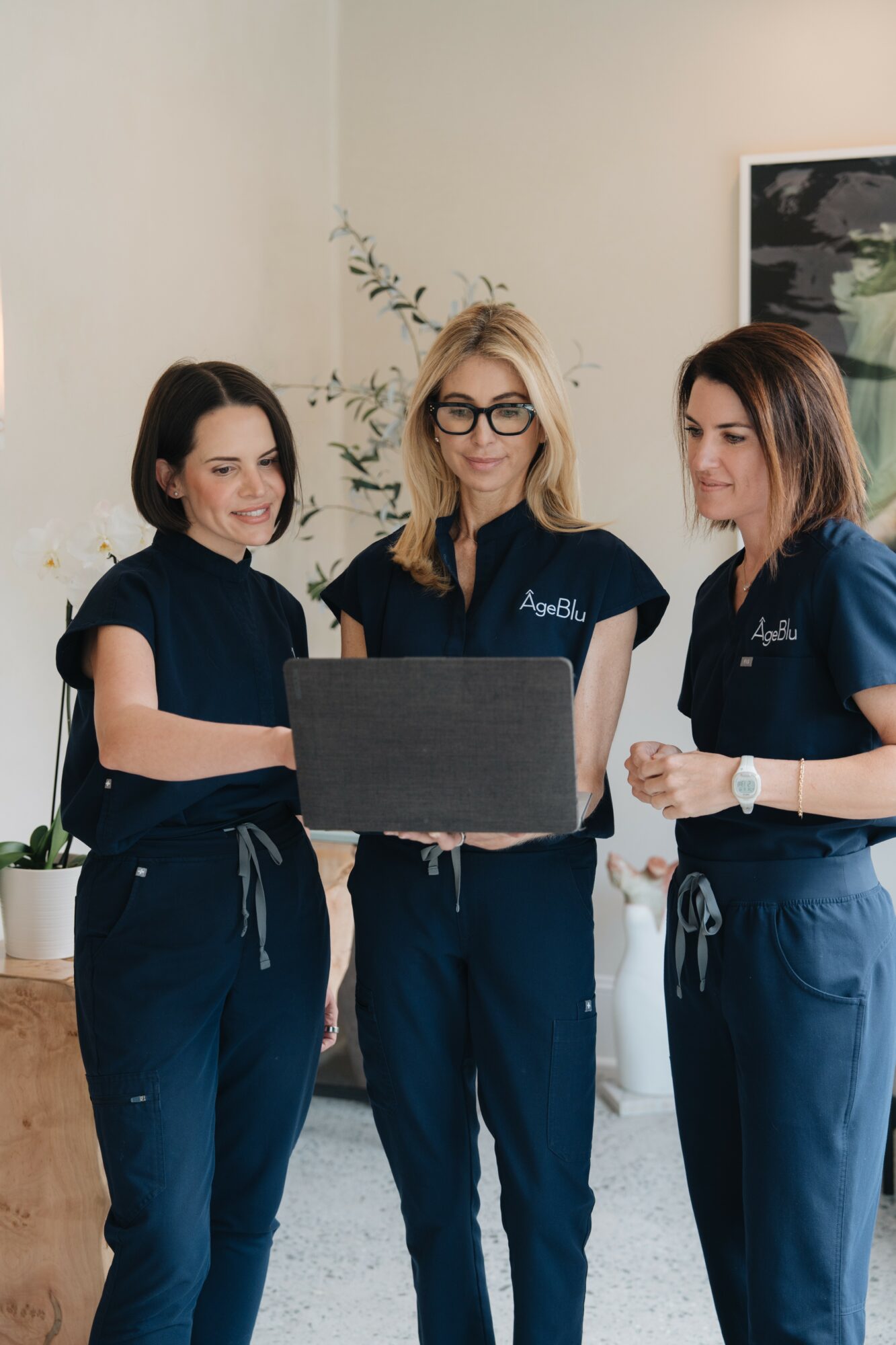 Three women in navy scrubs looking at a laptop together in a room with a plant and artwork