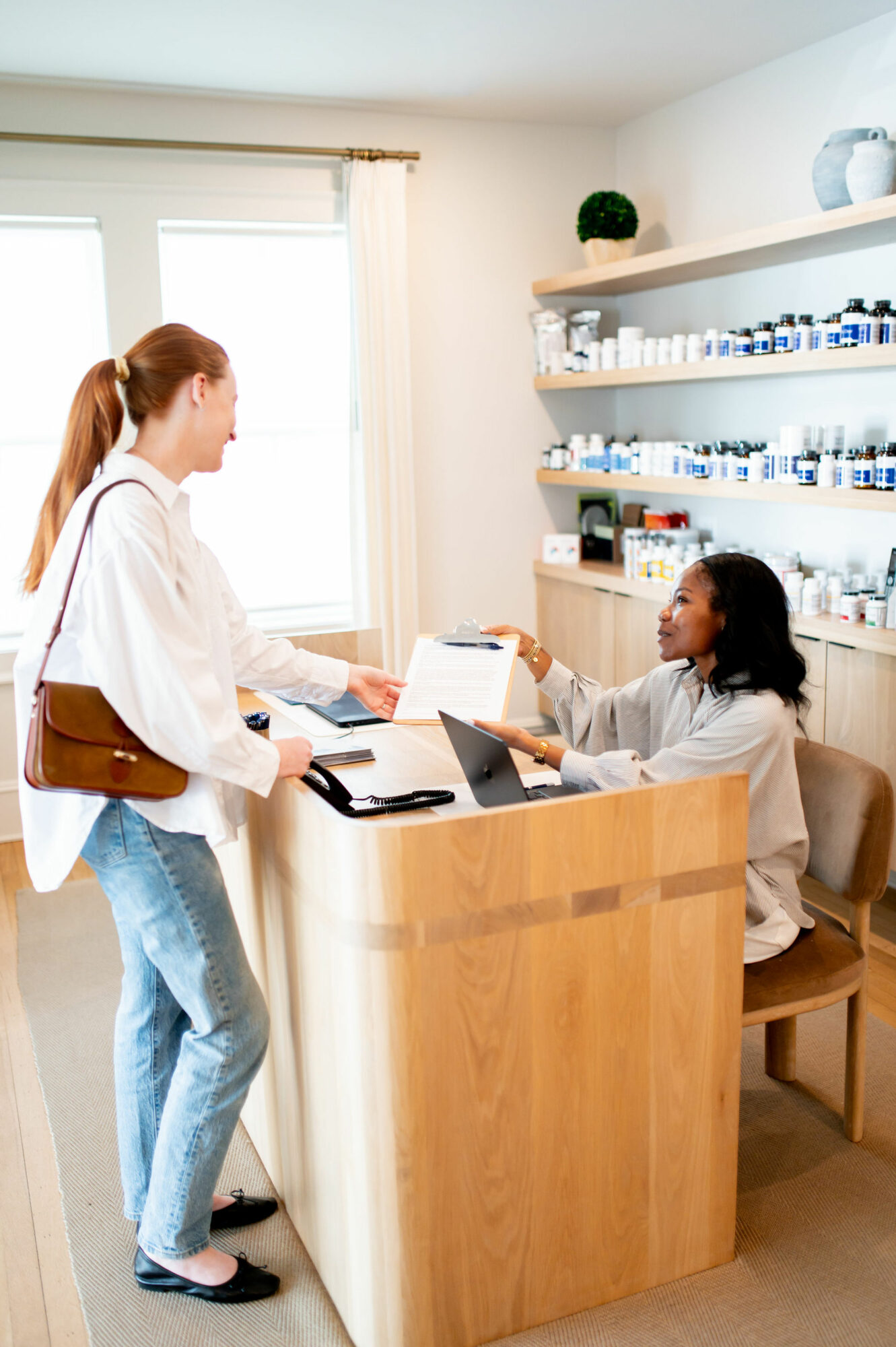 Two women at a reception desk in a bright, well-lit room with shelves of bottles and a window.