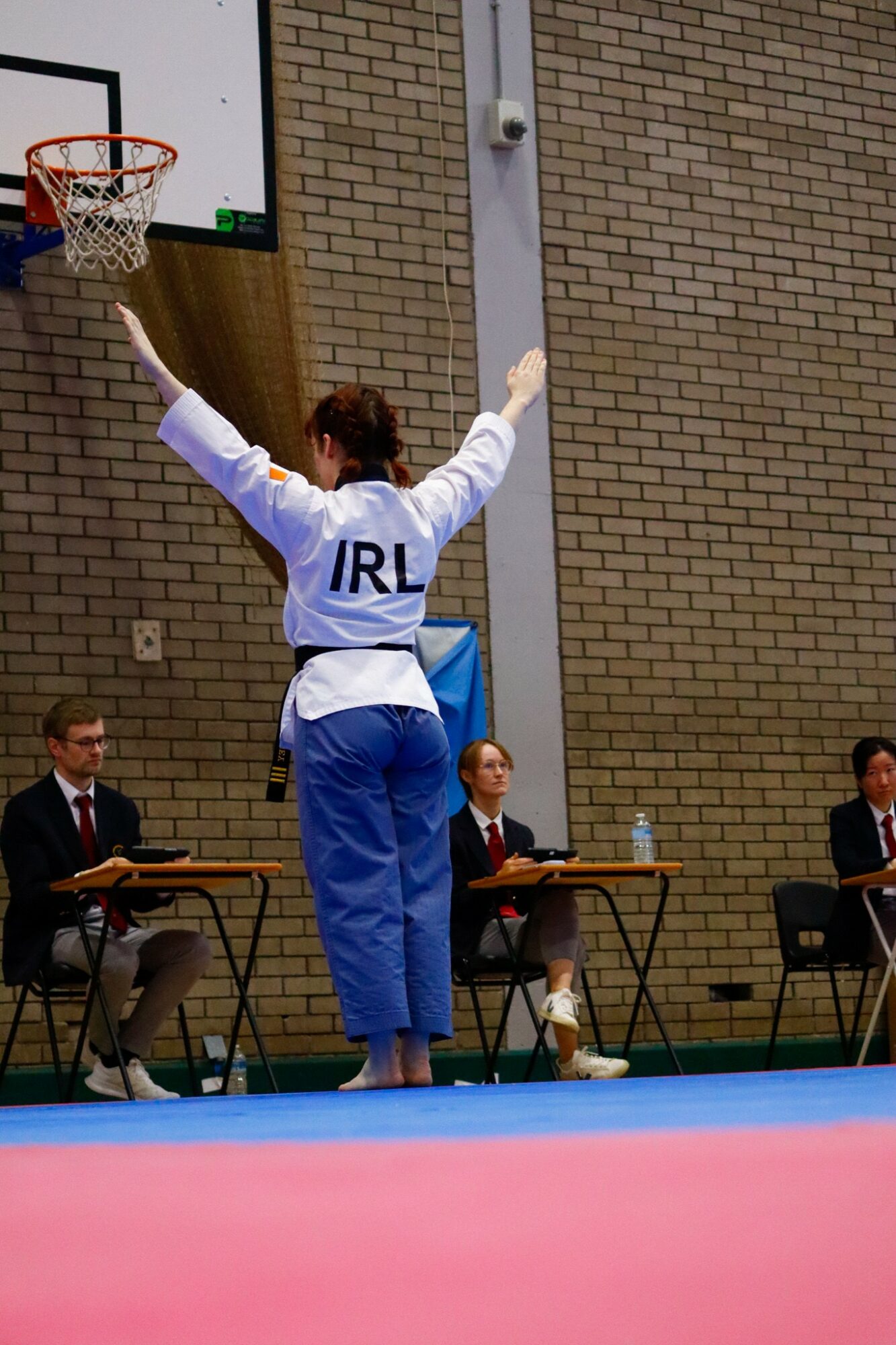 Person with arms raised, wearing a white and blue outfit with 'IRL' on the back, standing on a blue mat in a gymnasium.