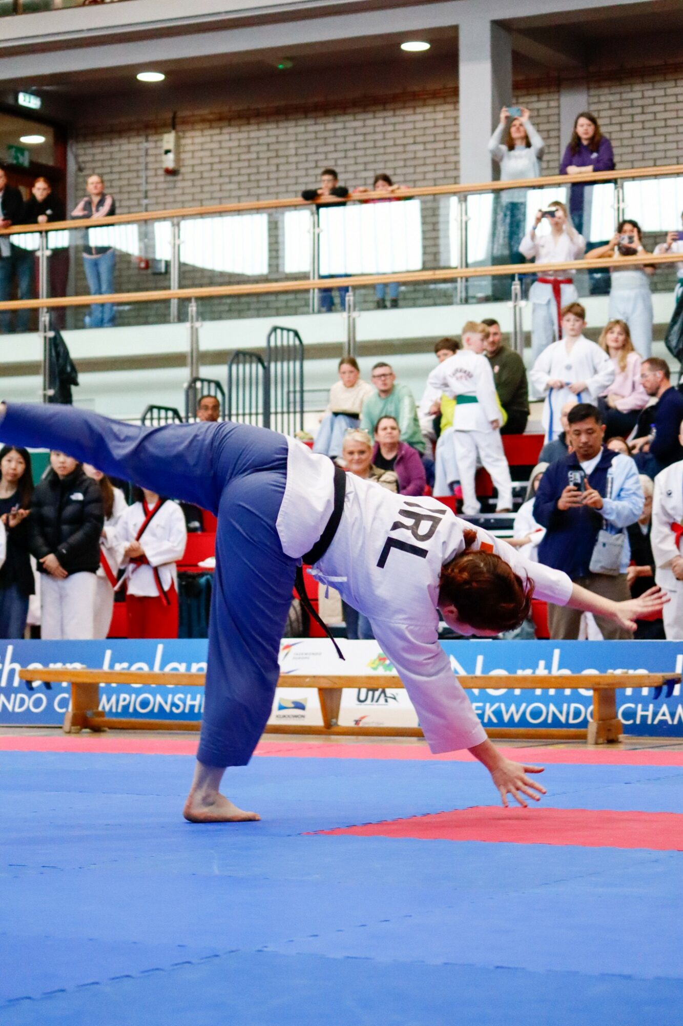 Person performing a gymnastics move on a blue mat with spectators watching from the background.