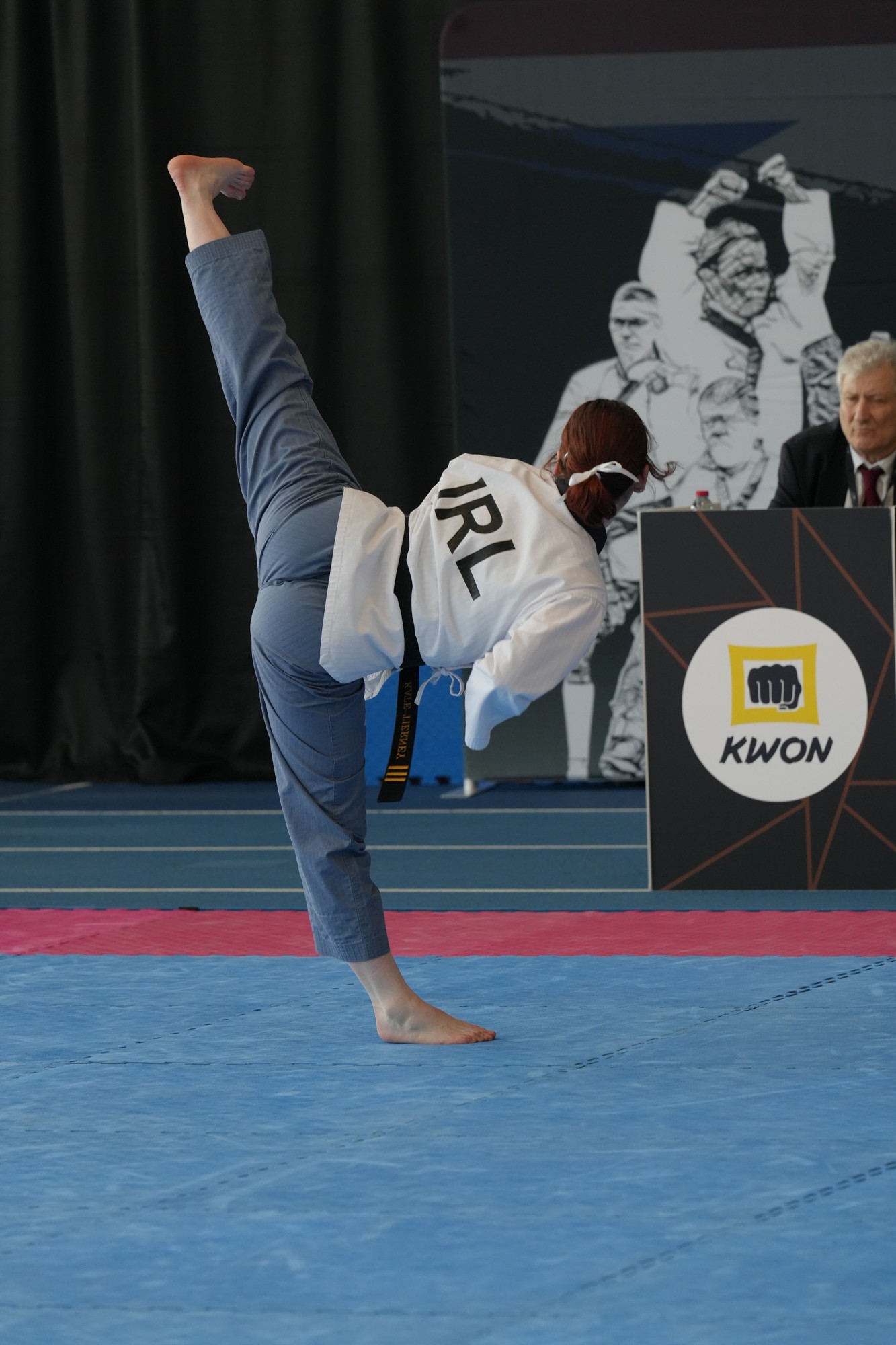 Person performing a handstand on a mat, wearing a white jacket with 'IRL' on the back, in a sports setting.