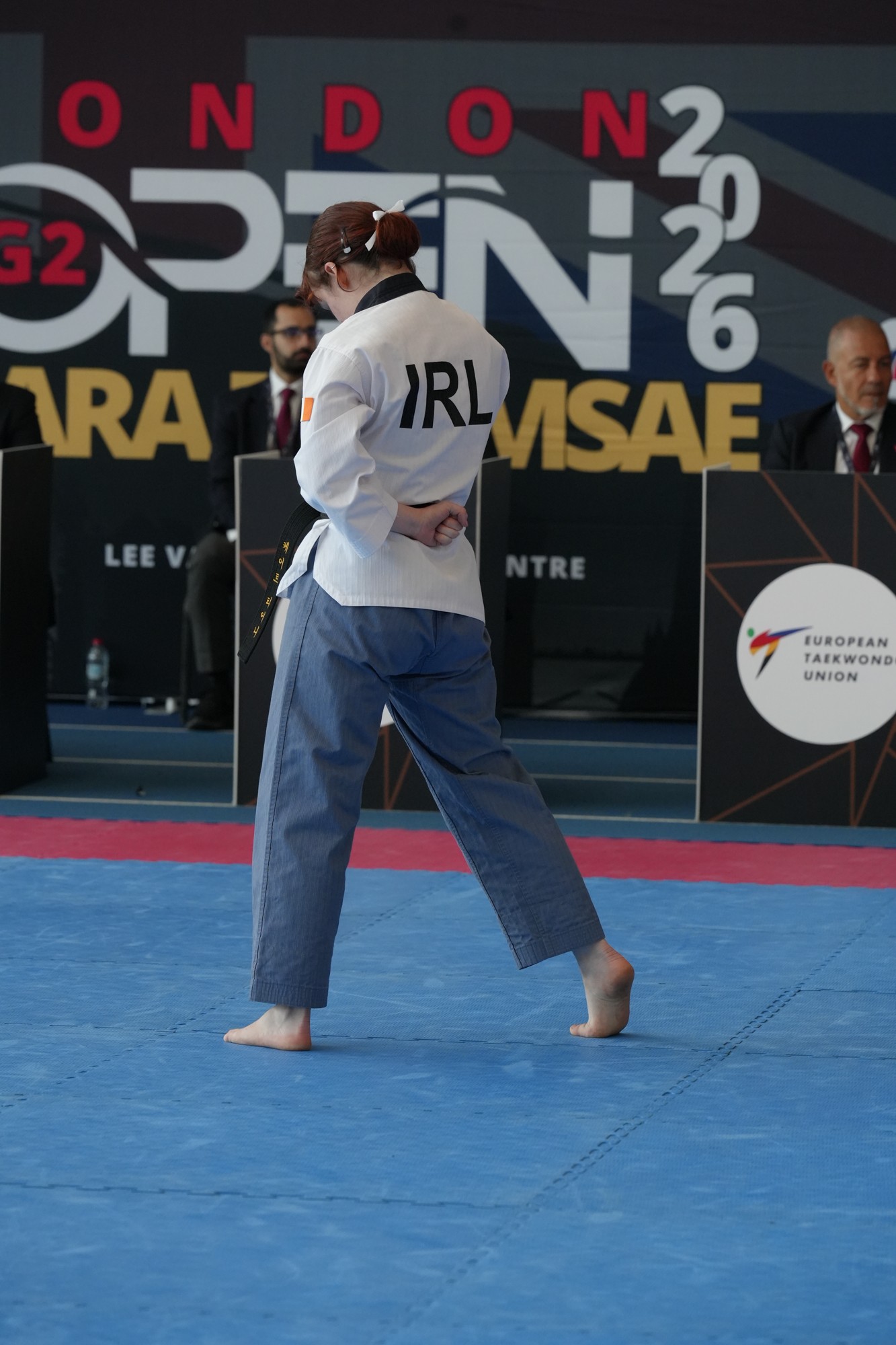 Person in martial arts uniform standing on blue mat with event banners in background.