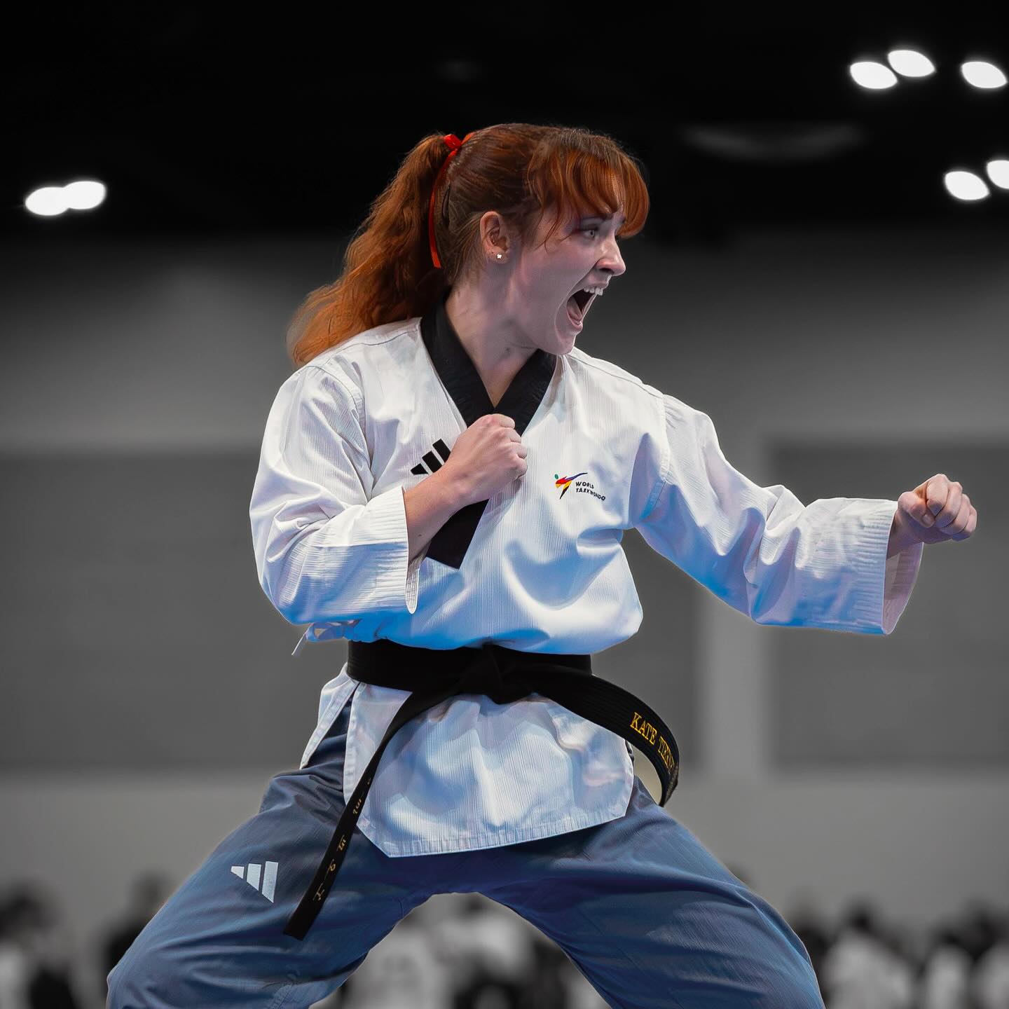 Female martial artist in a white uniform with a black belt, shouting and punching in a fighting stance, indoor setting.