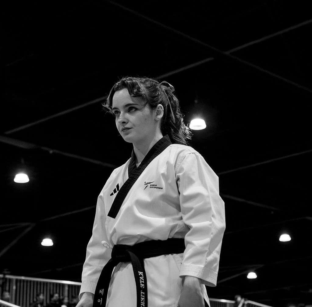 Young woman in martial arts uniform with black belt, standing indoors with ceiling lights visible.