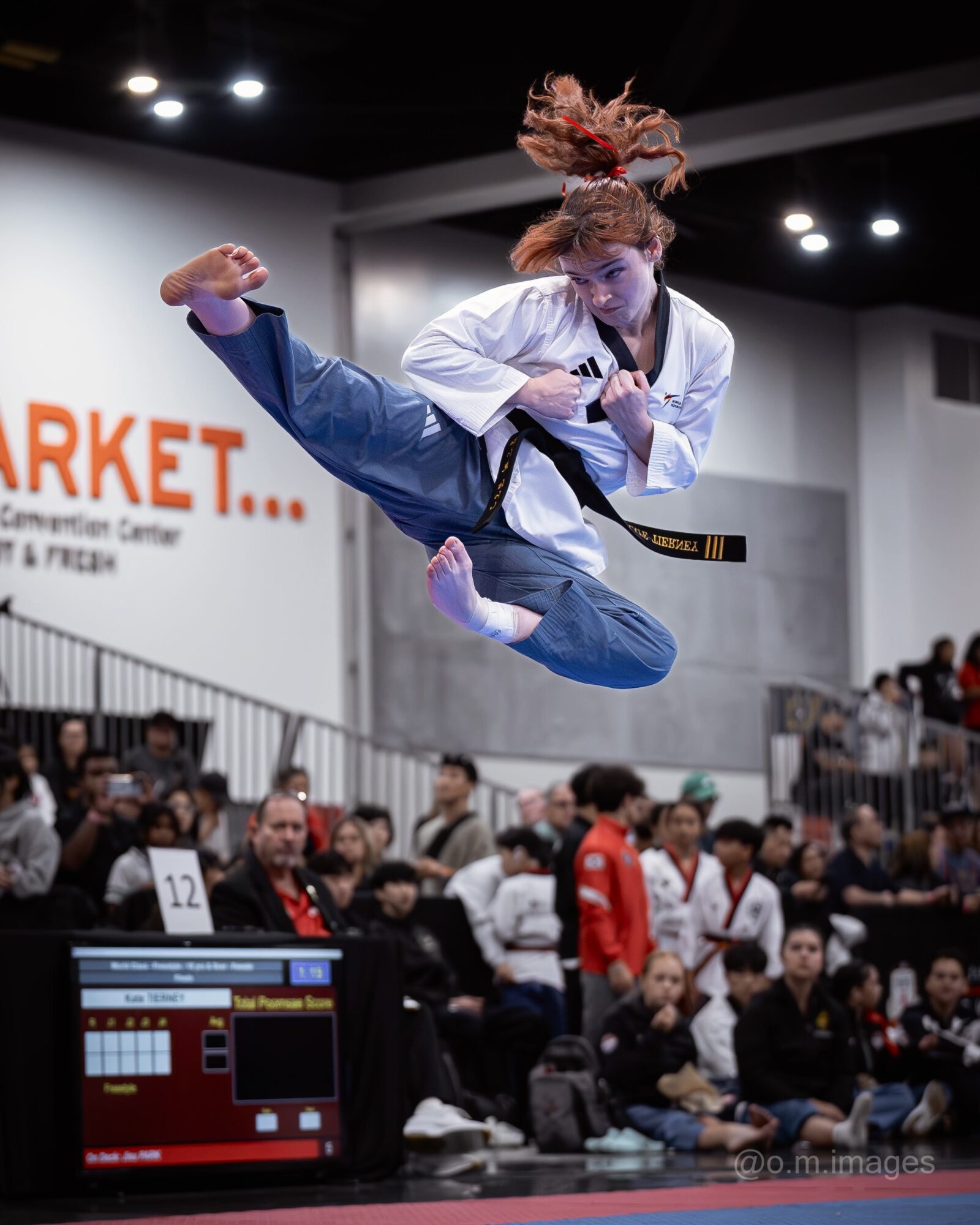 Martial artist in white gi mid-air performing a kick during a competition, audience watching below.