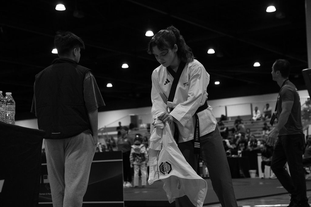A woman in a martial arts uniform adjusts her belt while two men watch in a gymnasium with spectators.