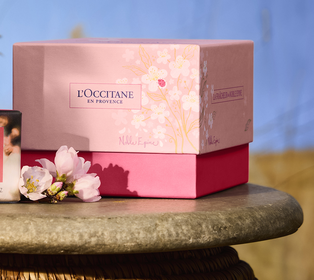 Pink and red L'Occitane gift boxes on a wooden surface with flowers nearby, against a blue background.