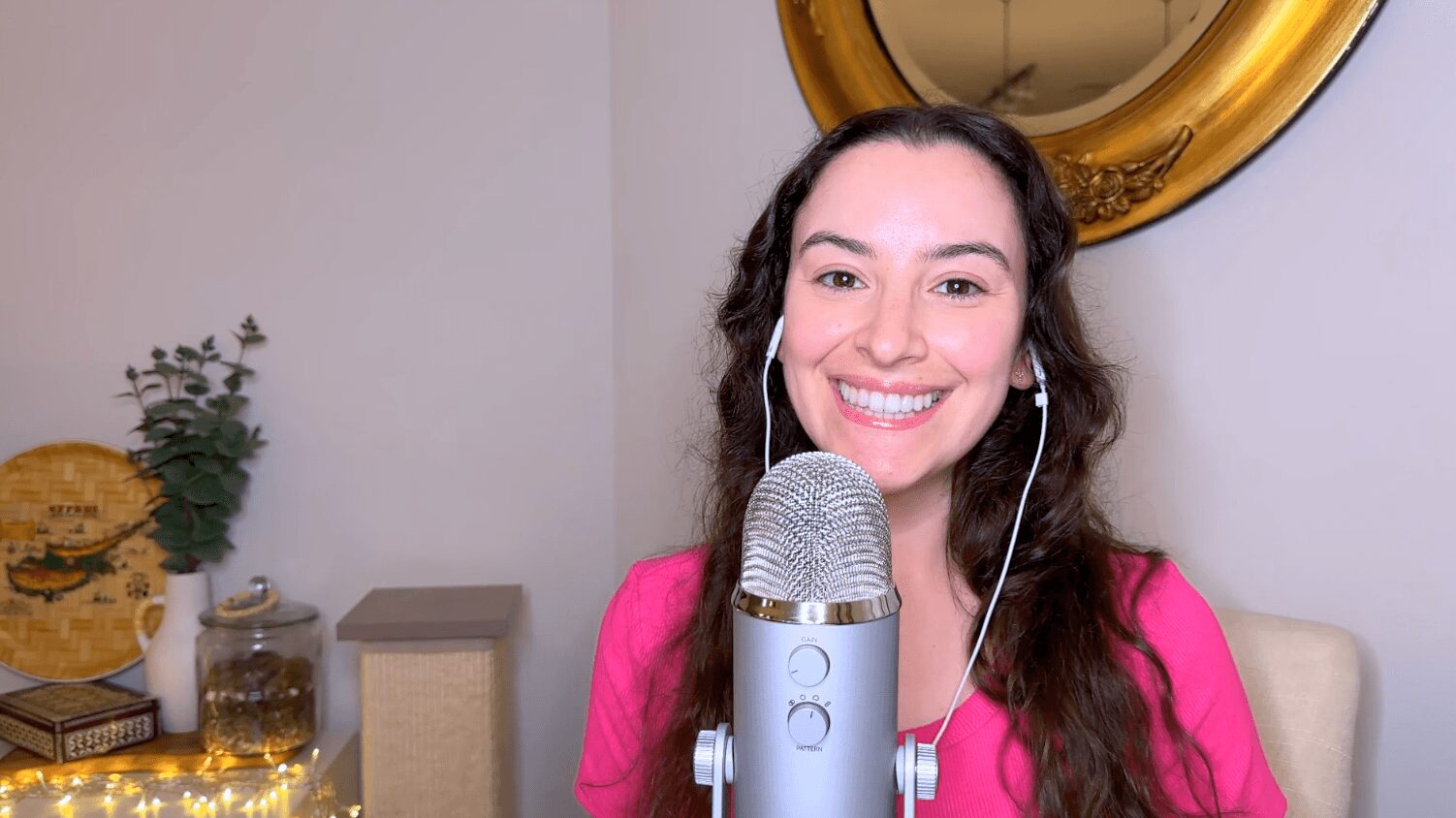 Young woman with long dark hair smiling, wearing pink, in front of a microphone, with a mirror and plants in background.