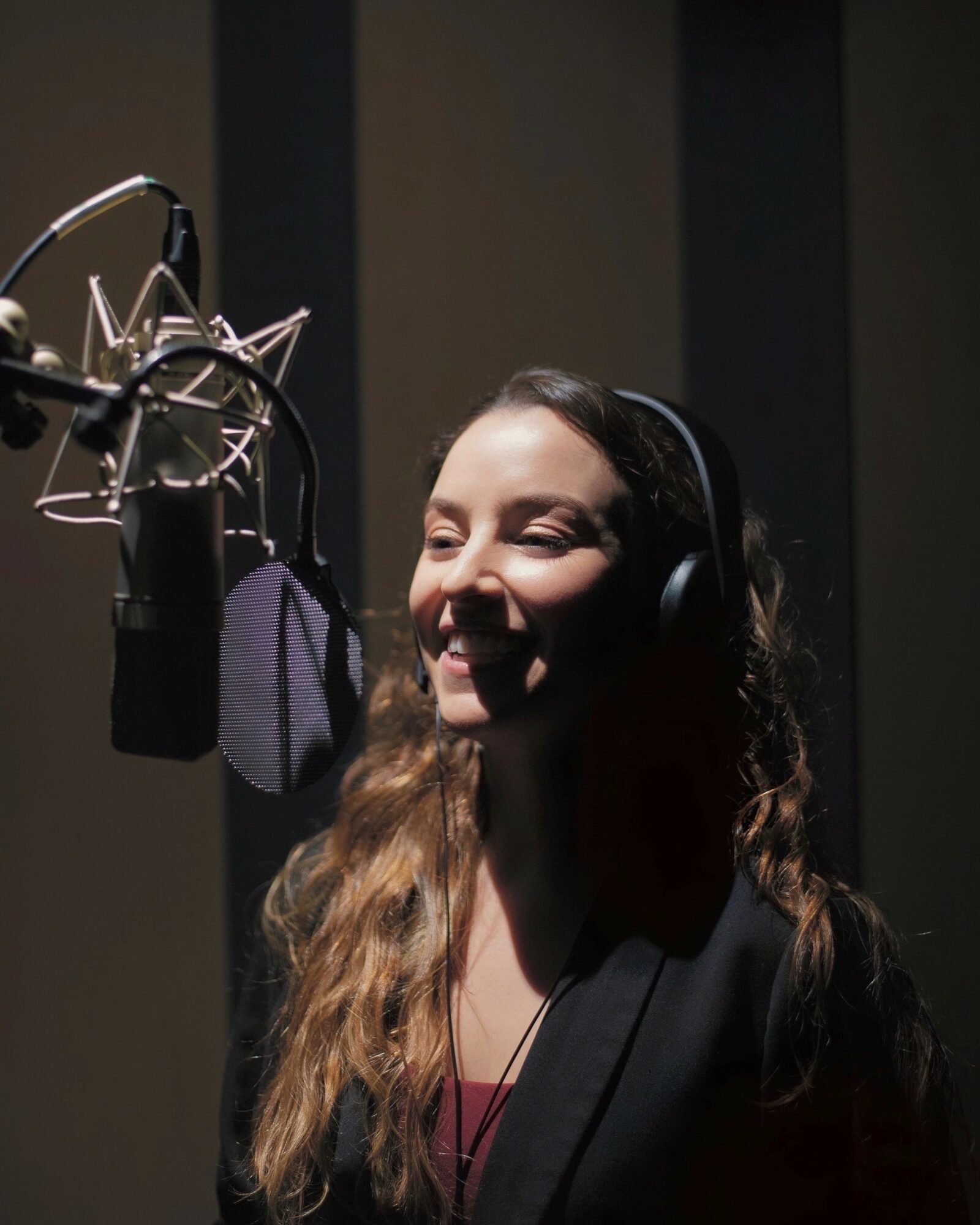 Woman with long hair wearing headphones, smiling, in a recording studio with a microphone.