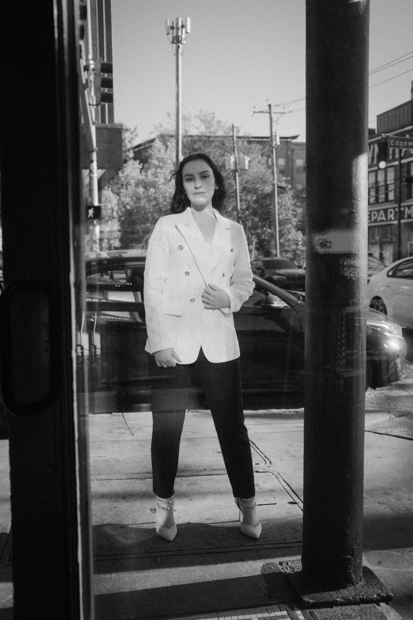 Woman standing on a city sidewalk, wearing a white blazer, black pants, and heels, with buildings and cars in background.