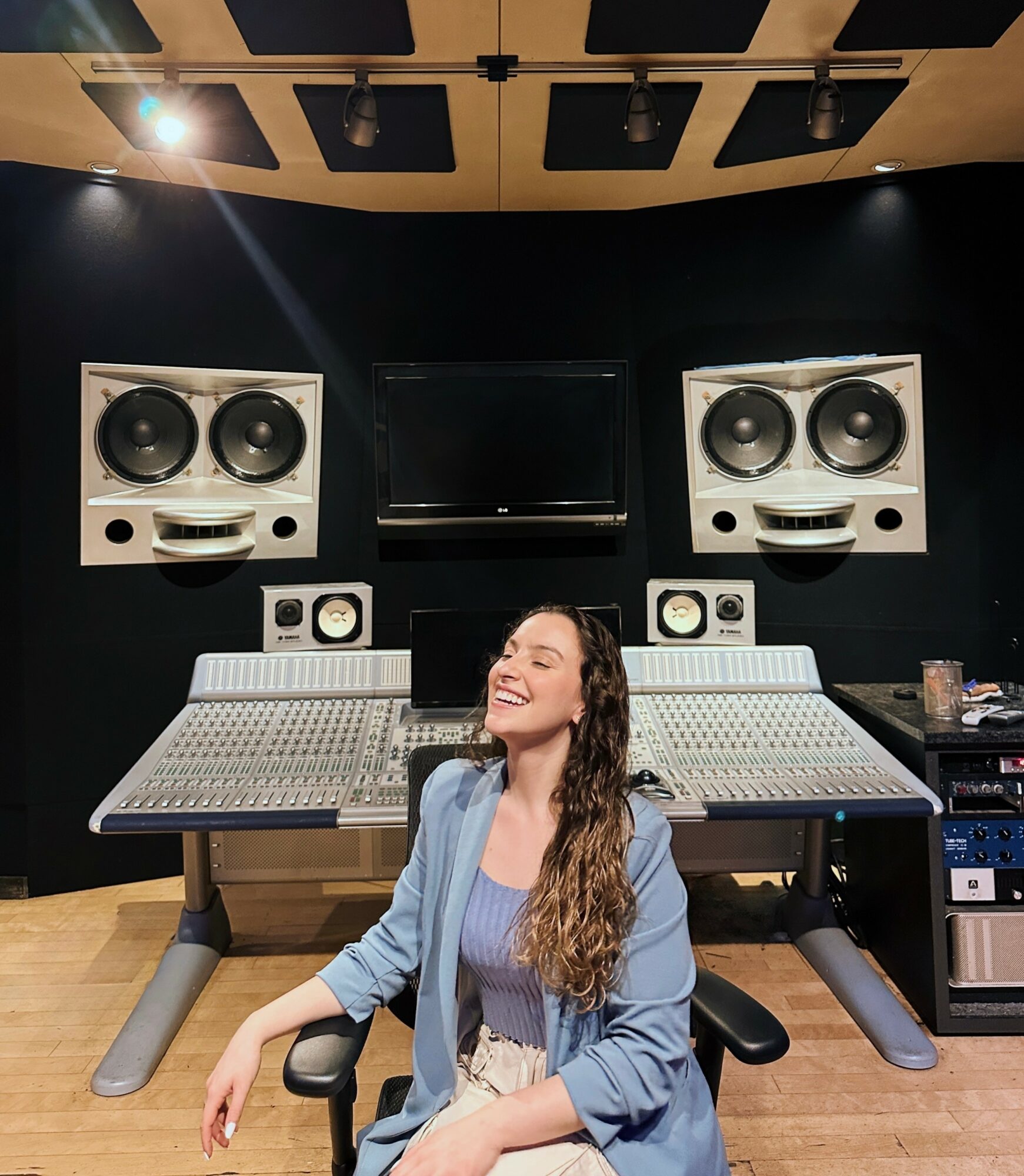 Young woman with long curly hair smiling in a recording studio with large speakers and audio equipment behind her.