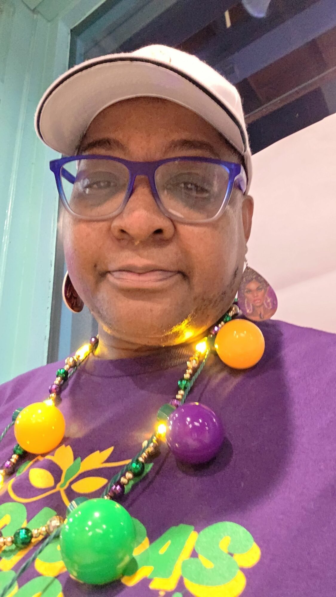 Person wearing glasses and a cap, smiling, with colorful beads and large beads around neck, in front of a purple background.