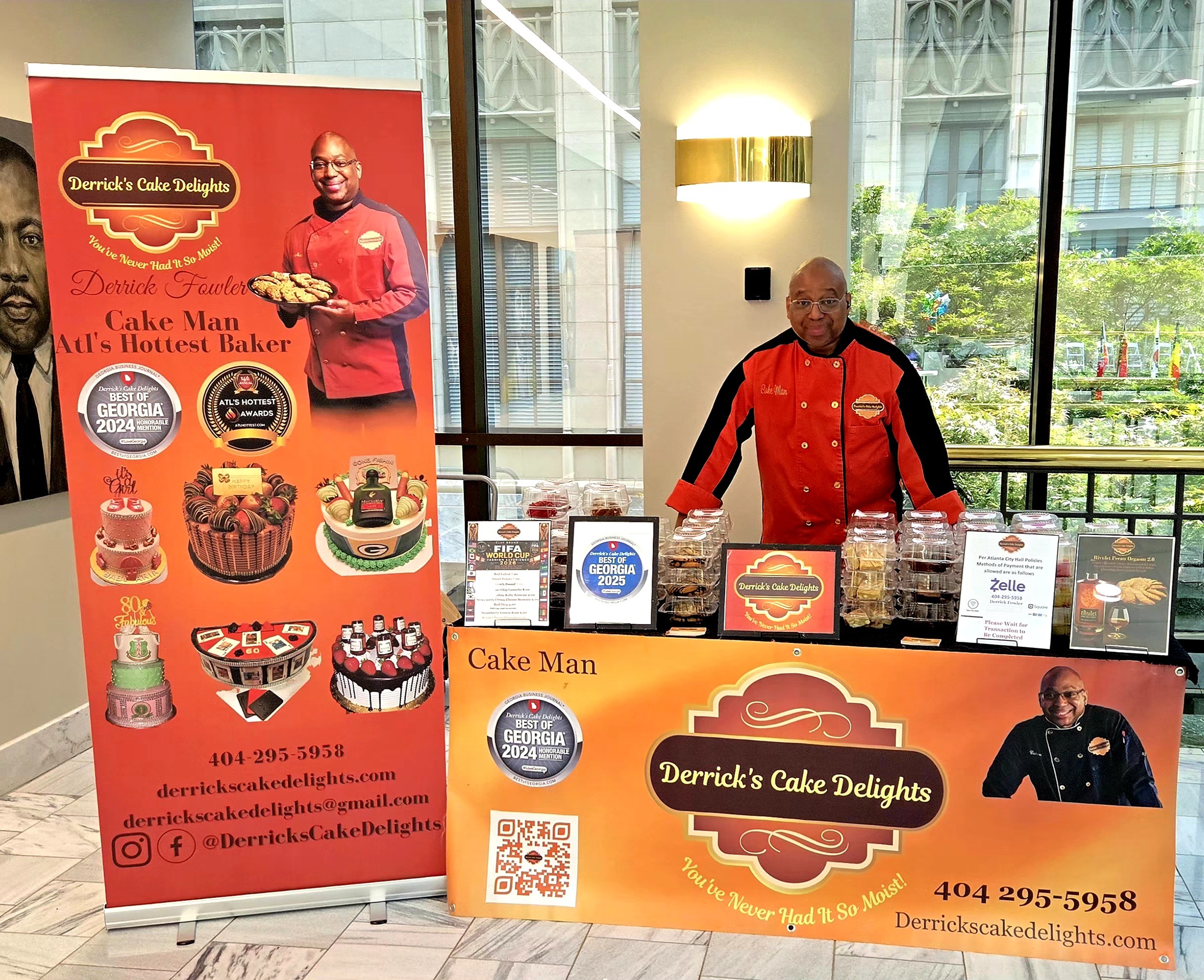 Man in red jacket standing behind cake display table with promotional banners, large windows in background, and indoor lighting.