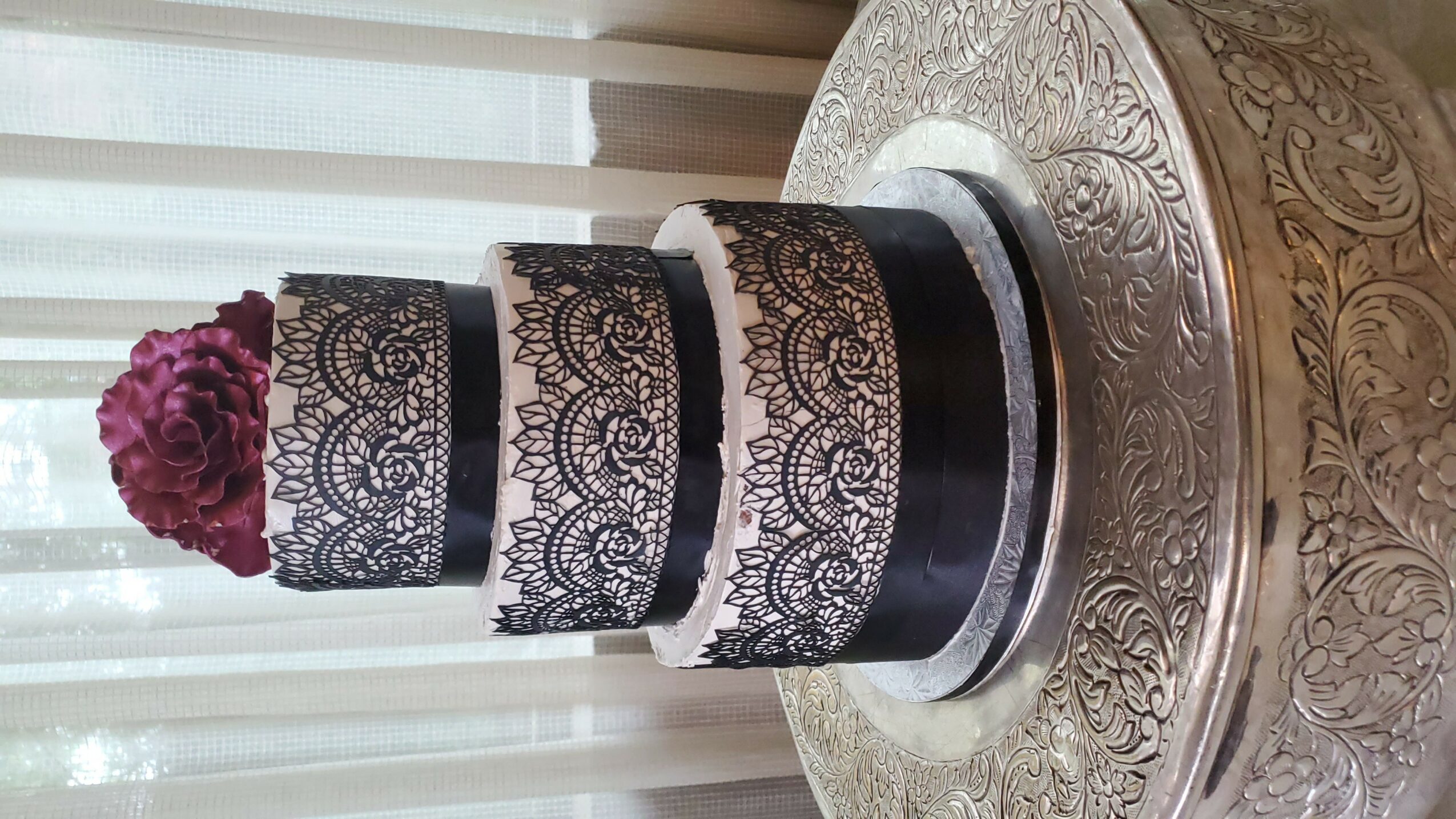 Multi-tiered cake with lace-like black patterns and a purple flower on top, placed on a round table with a decorative surface.