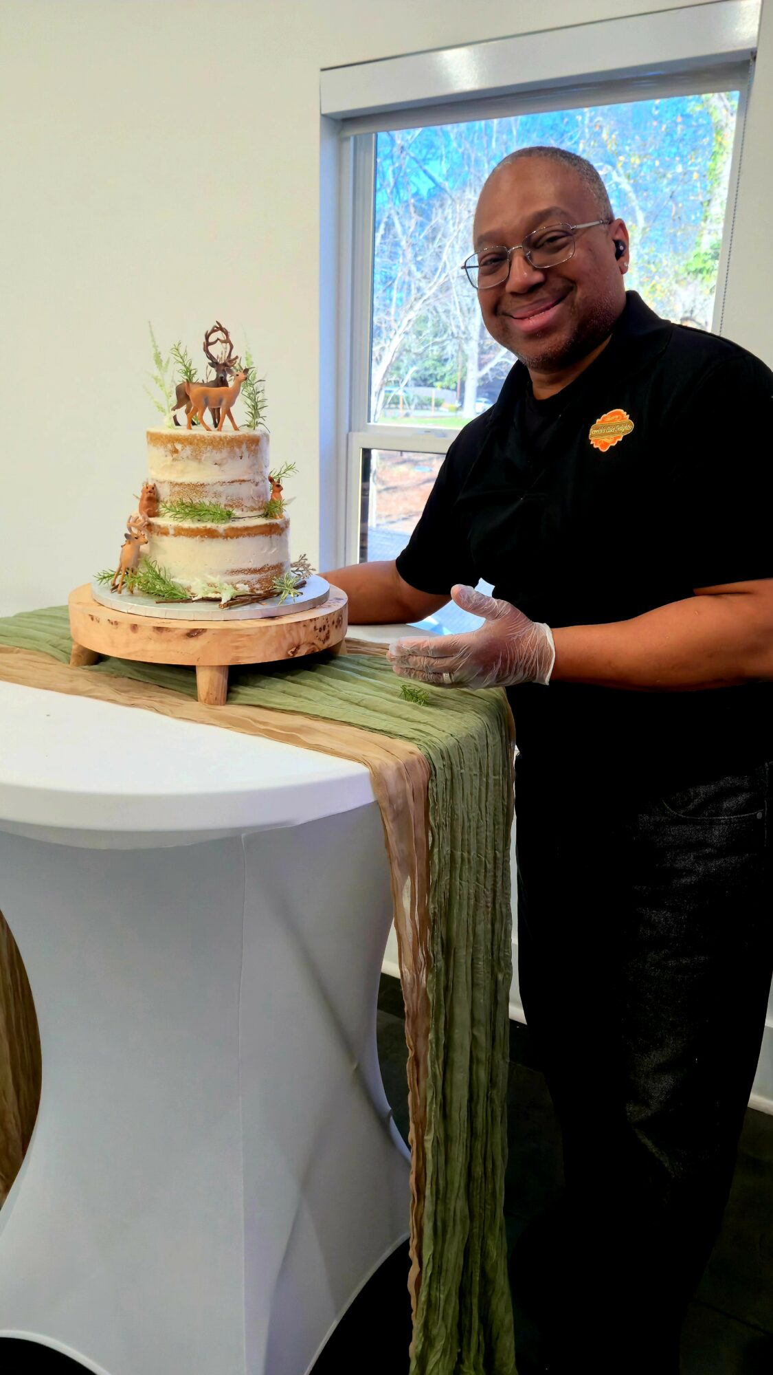Person smiling with a cake on a table, window with trees in background, wearing black shirt, holding a cake server.