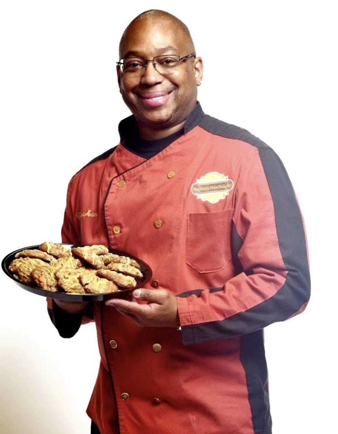 Man in chef uniform holding a tray of cookies, smiling, against a white background.