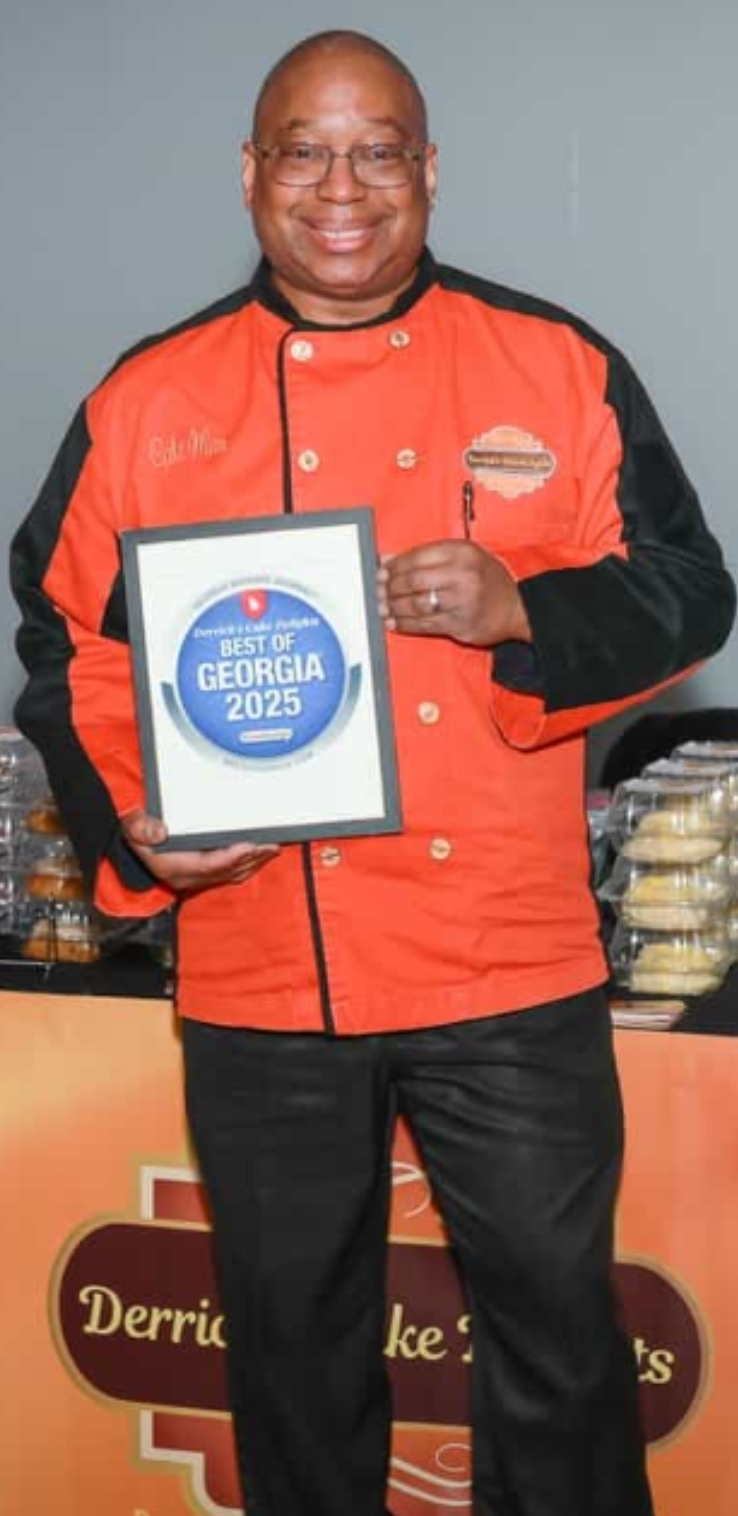 Man in red and black chef's jacket holding a framed certificate, smiling, standing in front of a table with food items.