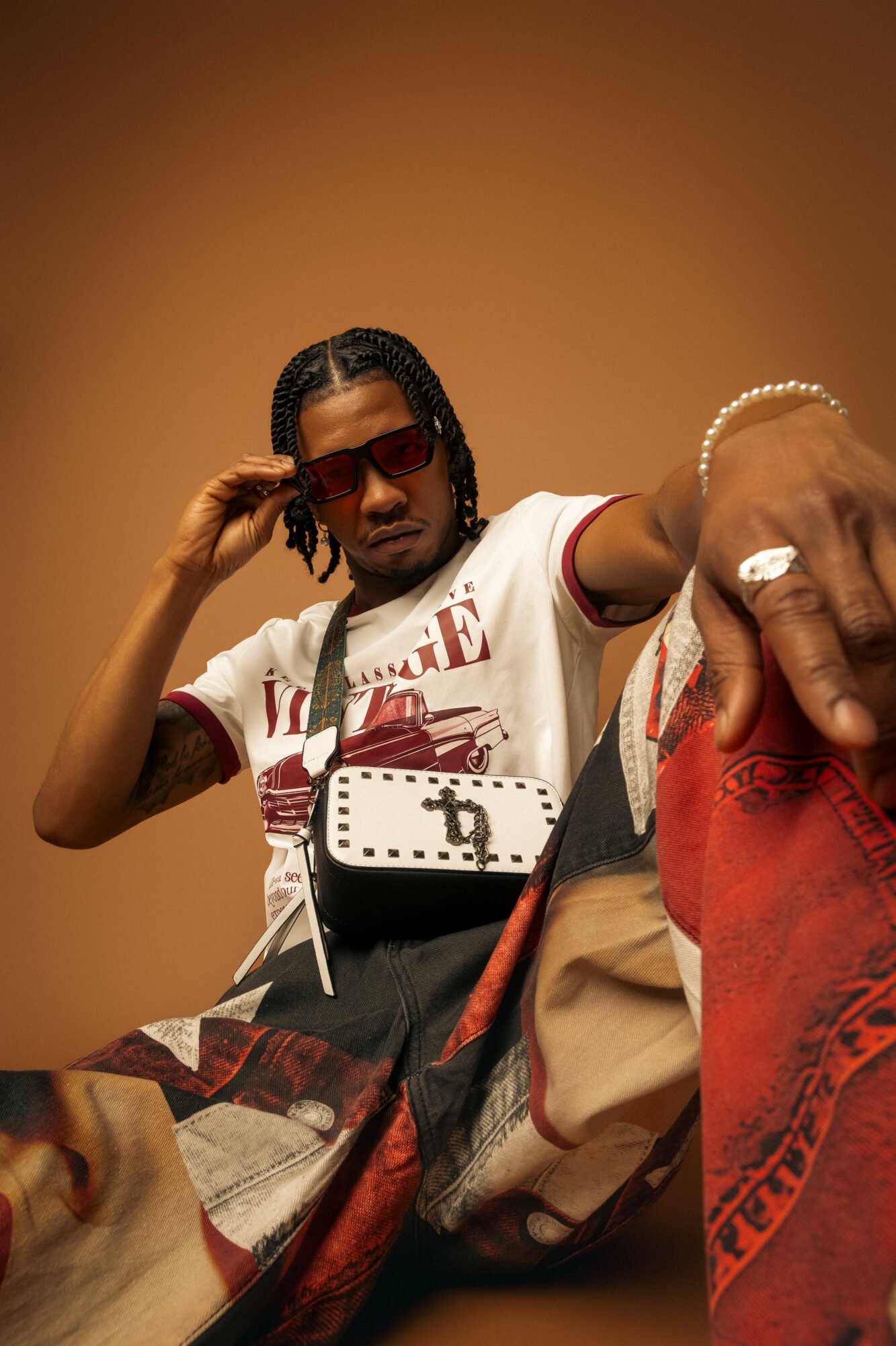 Person with braided hair wearing sunglasses, a white T-shirt, and jewelry, sitting against a plain background.