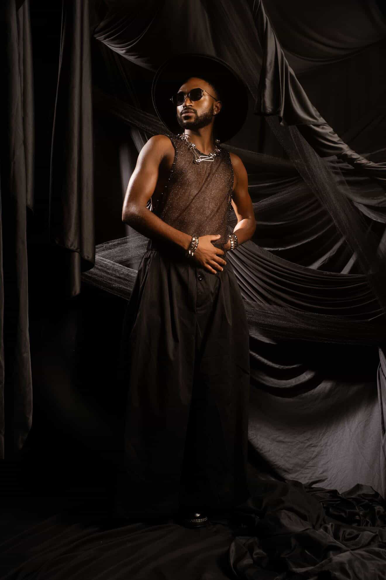 Person with sunglasses and jewelry stands against black draped fabric backdrop, wearing a sleeveless top and long skirt.