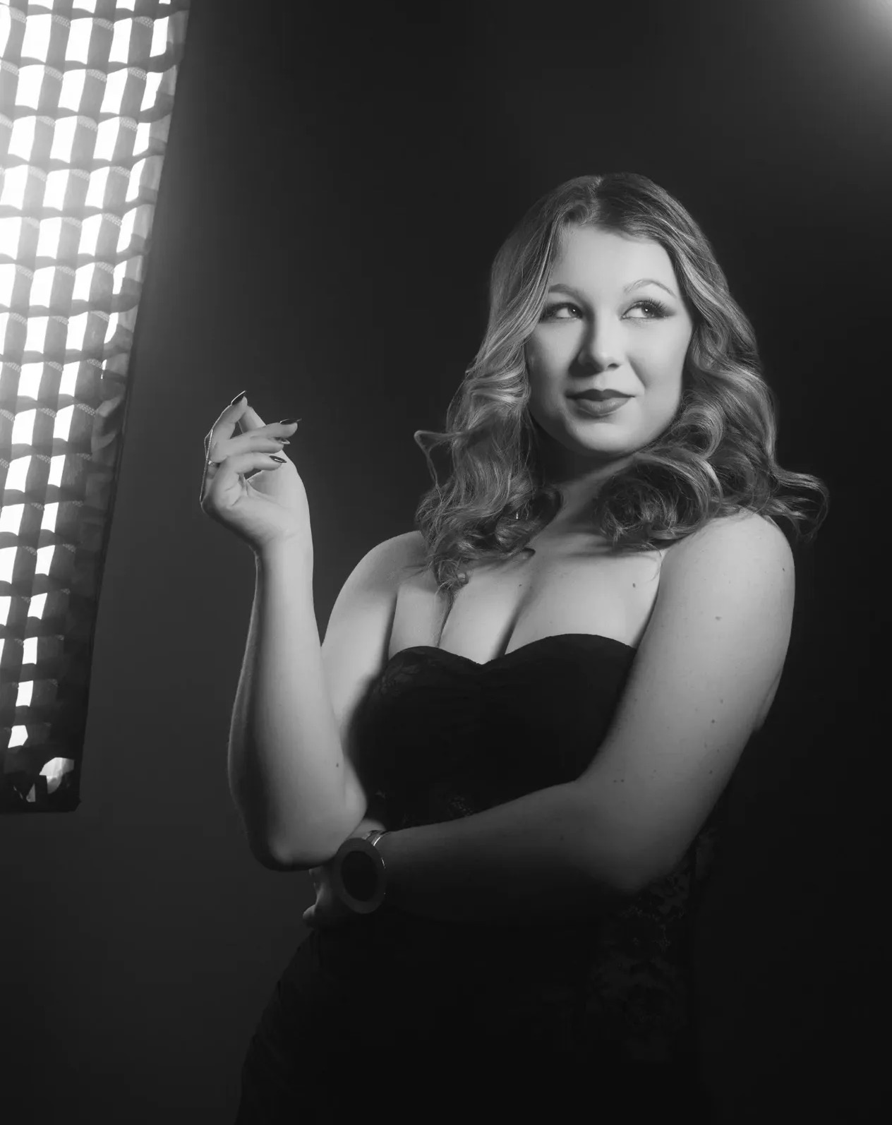 A woman with wavy hair in a strapless dress poses with her hand near her face, smiling slightly, in a studio setting.