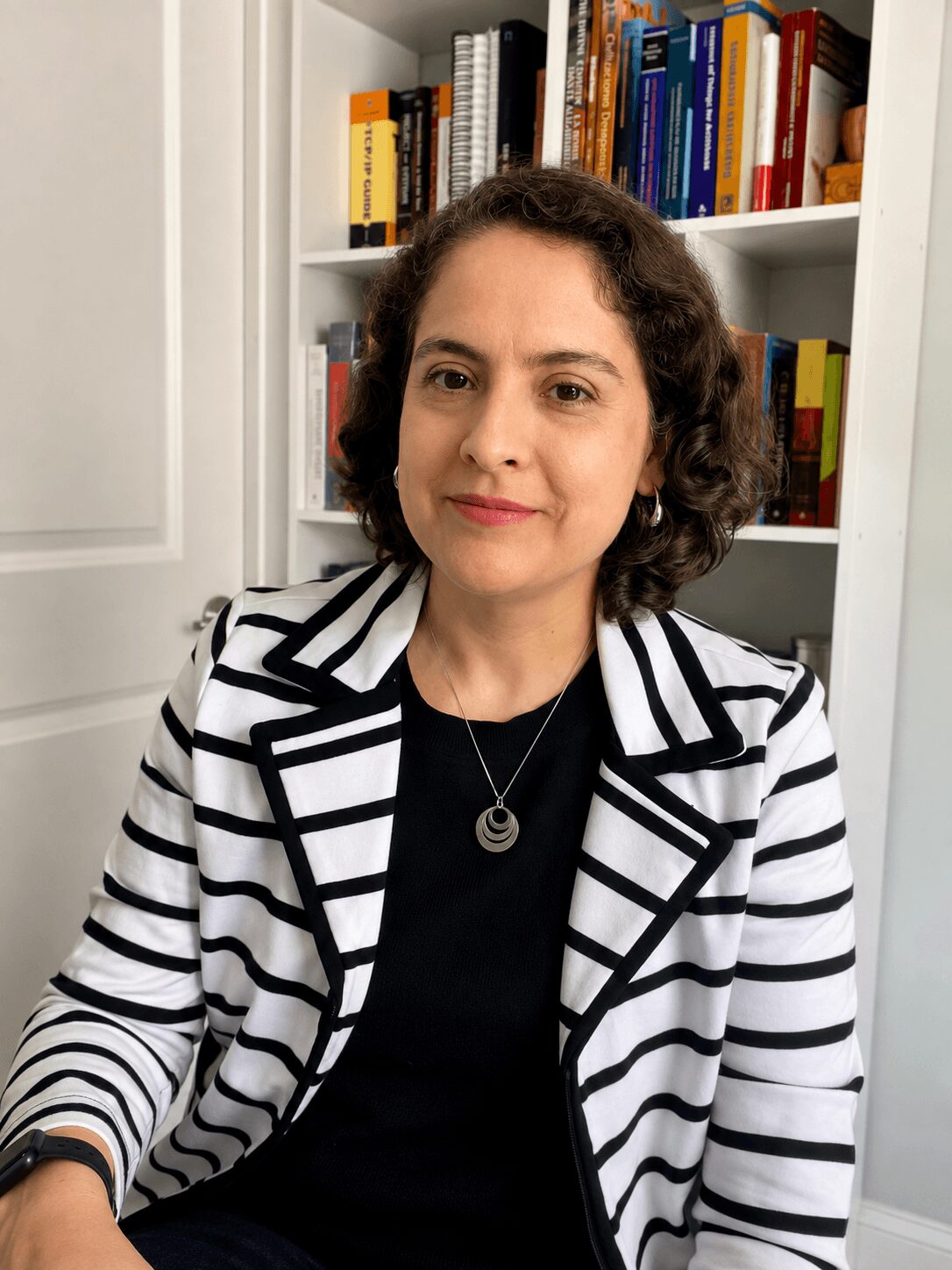 Woman with curly hair wearing a striped blazer and black top, sitting in front of a bookshelf filled with books.