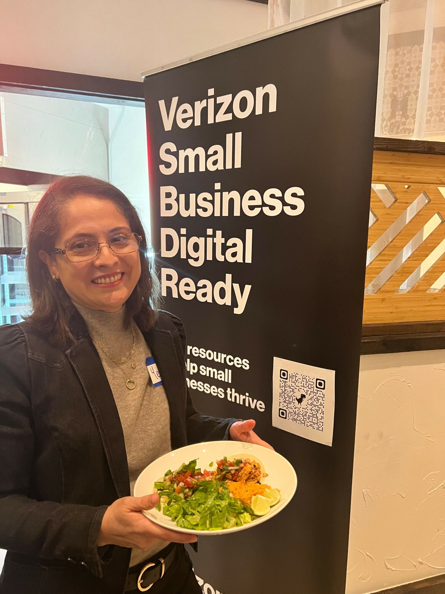 Smiling woman holding a plate of food, standing next to a black sign with white text about Verizon Small Business Digital Ready.