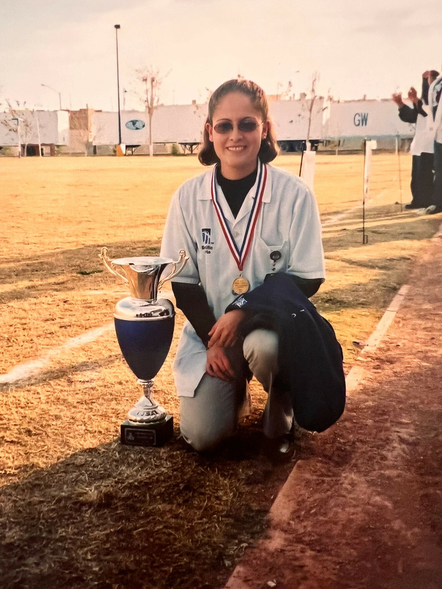 Person kneeling on a sports field with a trophy, wearing a medal and sports uniform, smiling at the camera.
