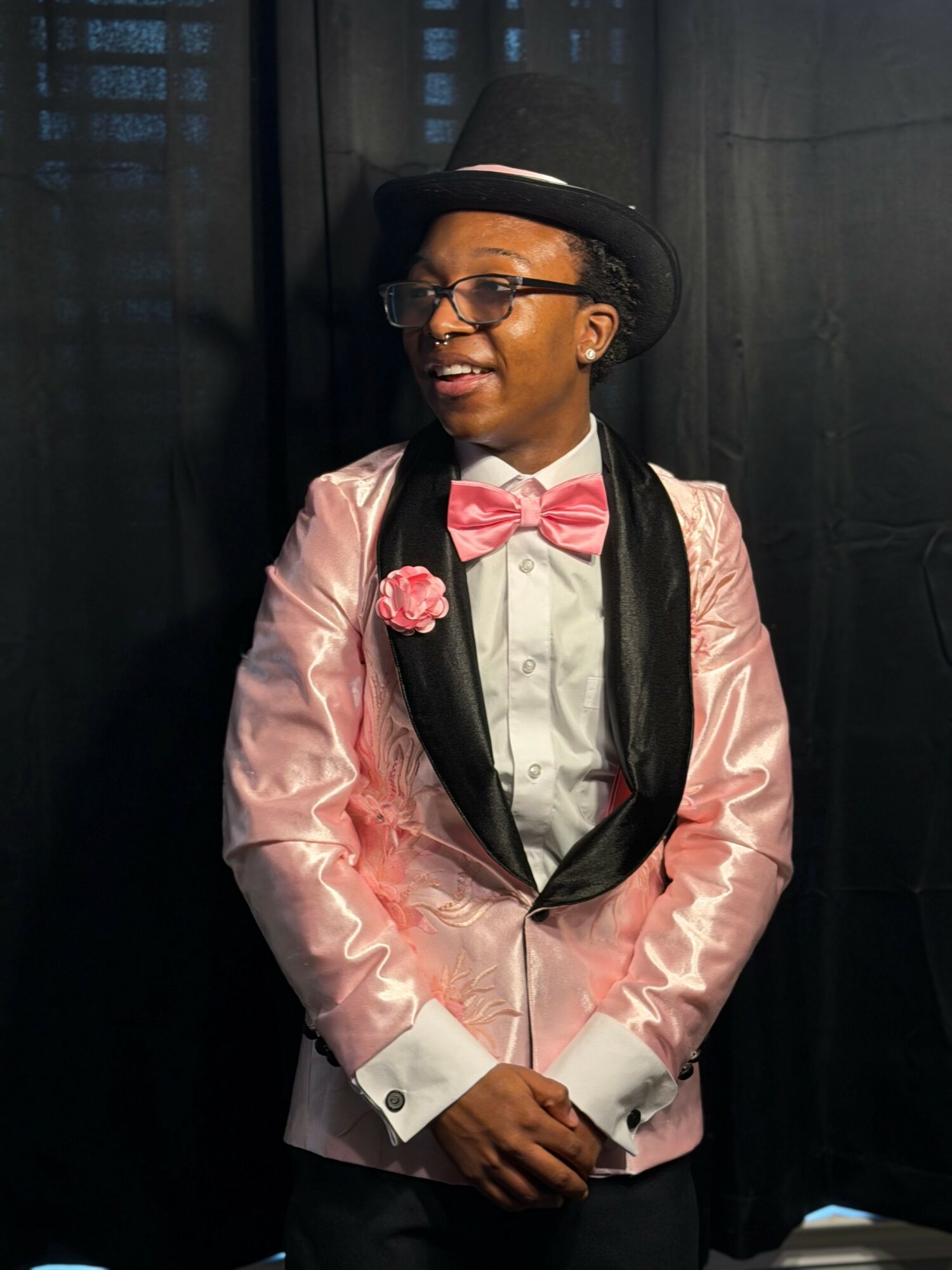 Young person wearing a pink satin jacket, pink bow tie, glasses, and a black top hat, smiling with hands clasped.