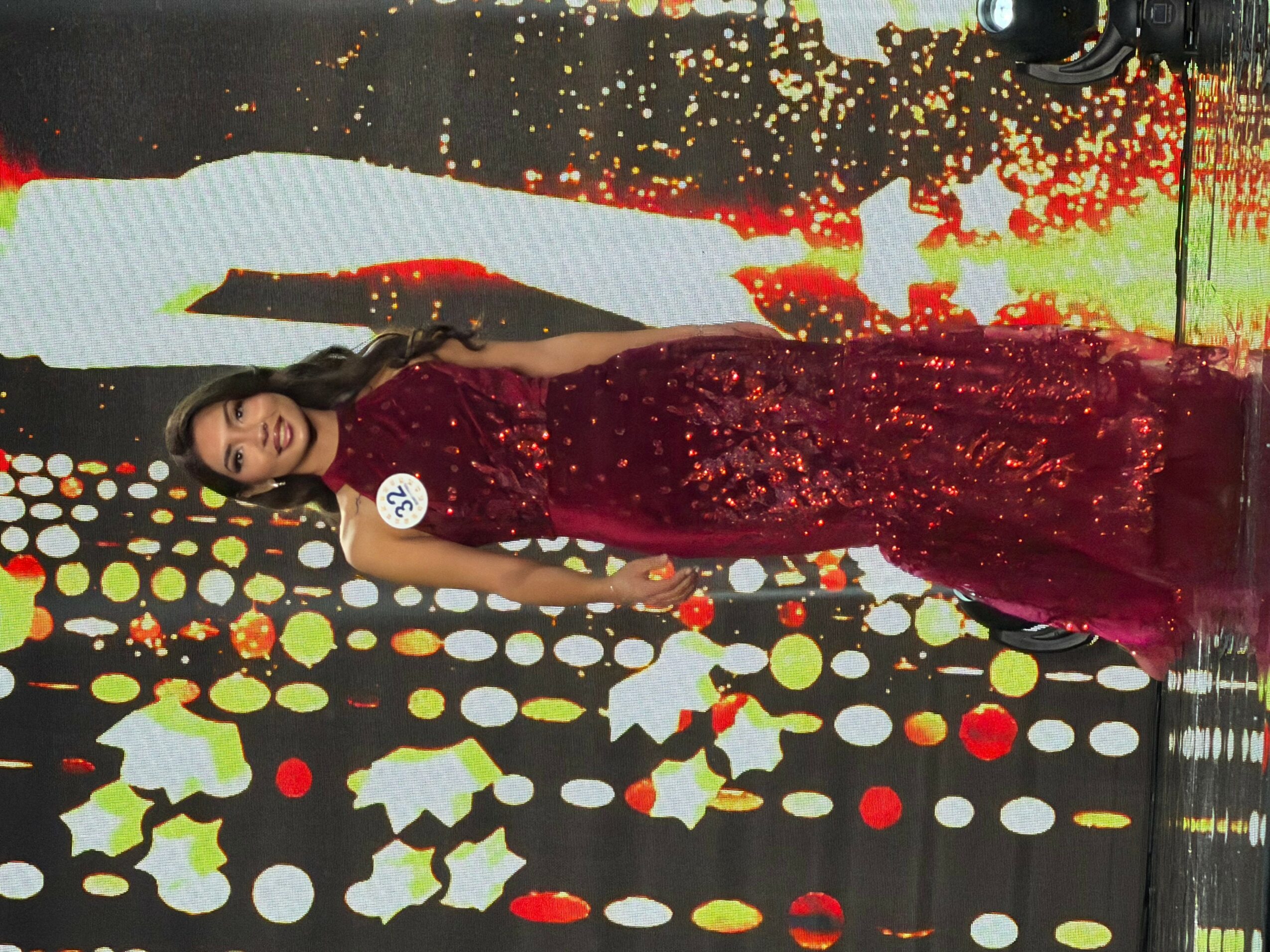 Woman in a red sequined gown standing on a colorful, dotted stage background, smiling and posing with her hands on her hips.