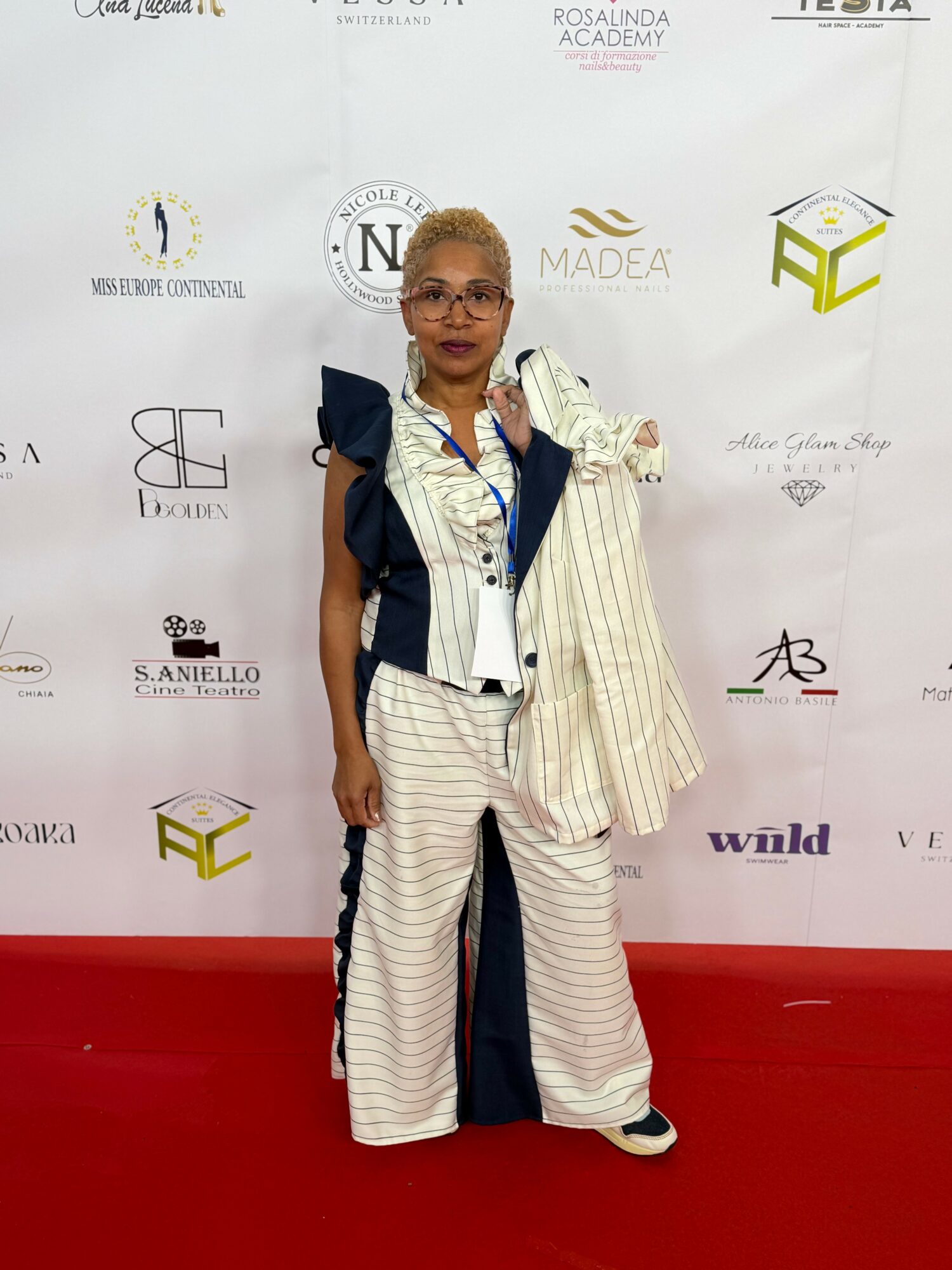 Woman with glasses and short hair in a striped outfit holding a bag, standing on a red carpet in front of a white backdrop.