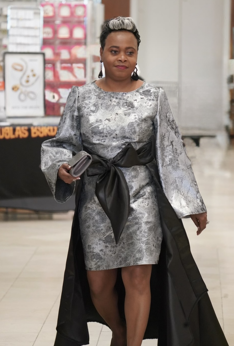 Woman in silver dress with black sash, holding a clutch, walking indoors in a shopping area.